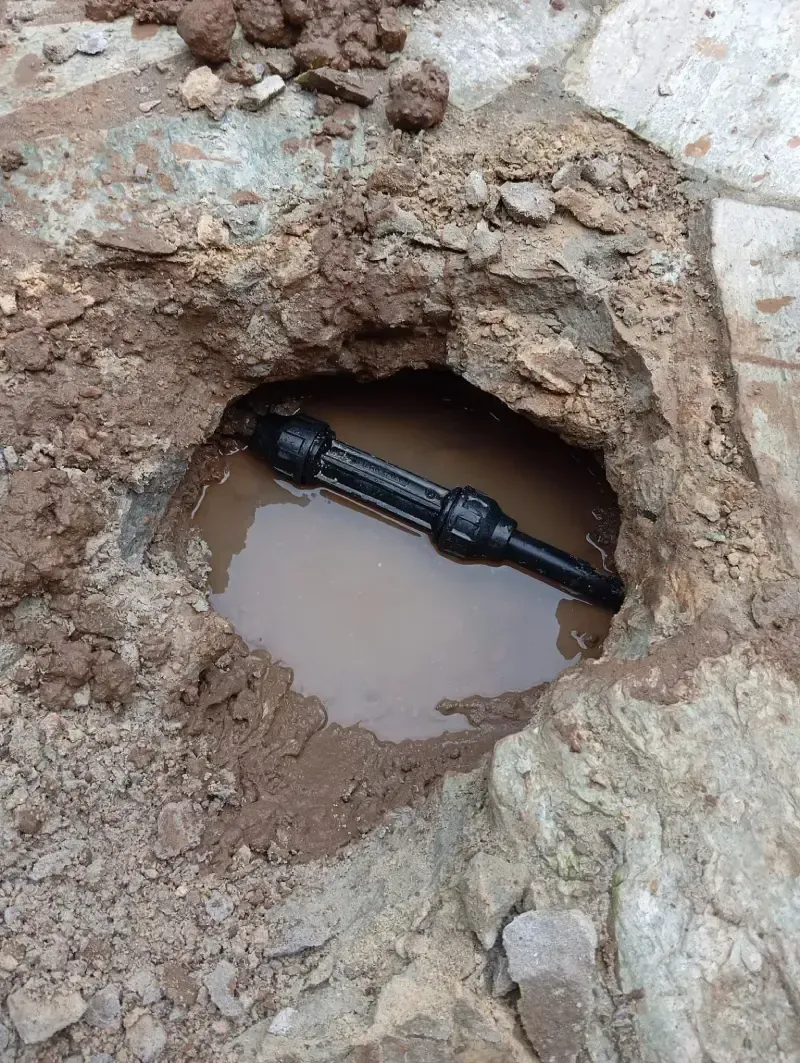 Un conector de tubería negro descansa en un pequeño agujero fangoso en el suelo, lleno de agua.