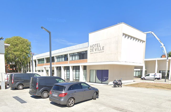 Extérieur de l'Hôtel de Ville en France avec des voitures garées par une journée ensoleillée.