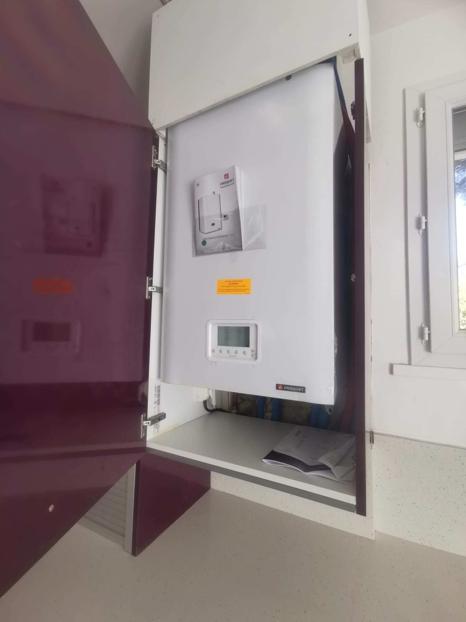 Une chaudière blanche est installée dans une armoire avec une porte marron partiellement ouverte, à l'intérieur d'une maison près d'une fenêtre.