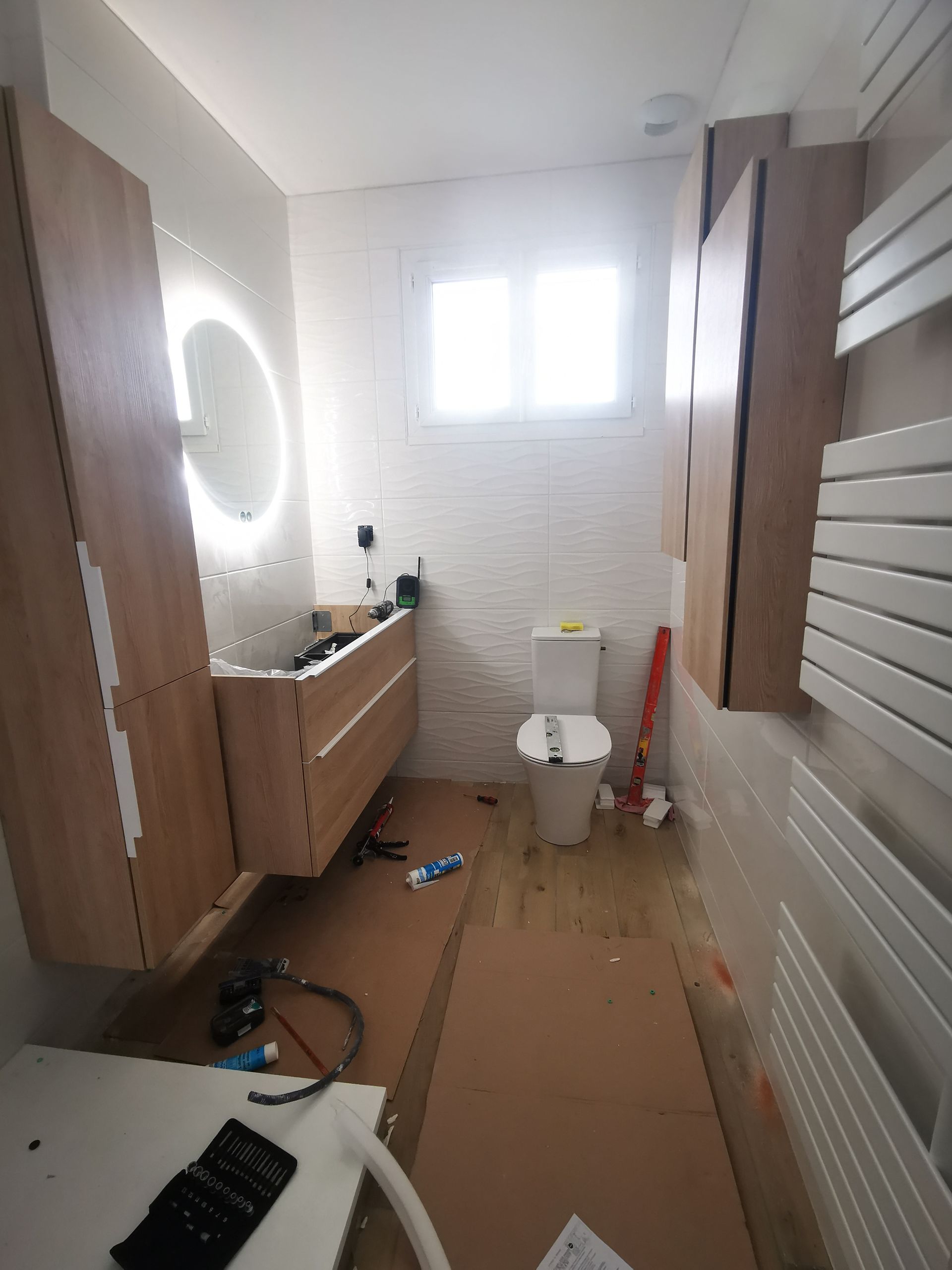 Rénovation de salle de bain : armoires en bois clair, toilette et fenêtre blanches, et un miroir avec lumière, entouré de matériaux de construction.