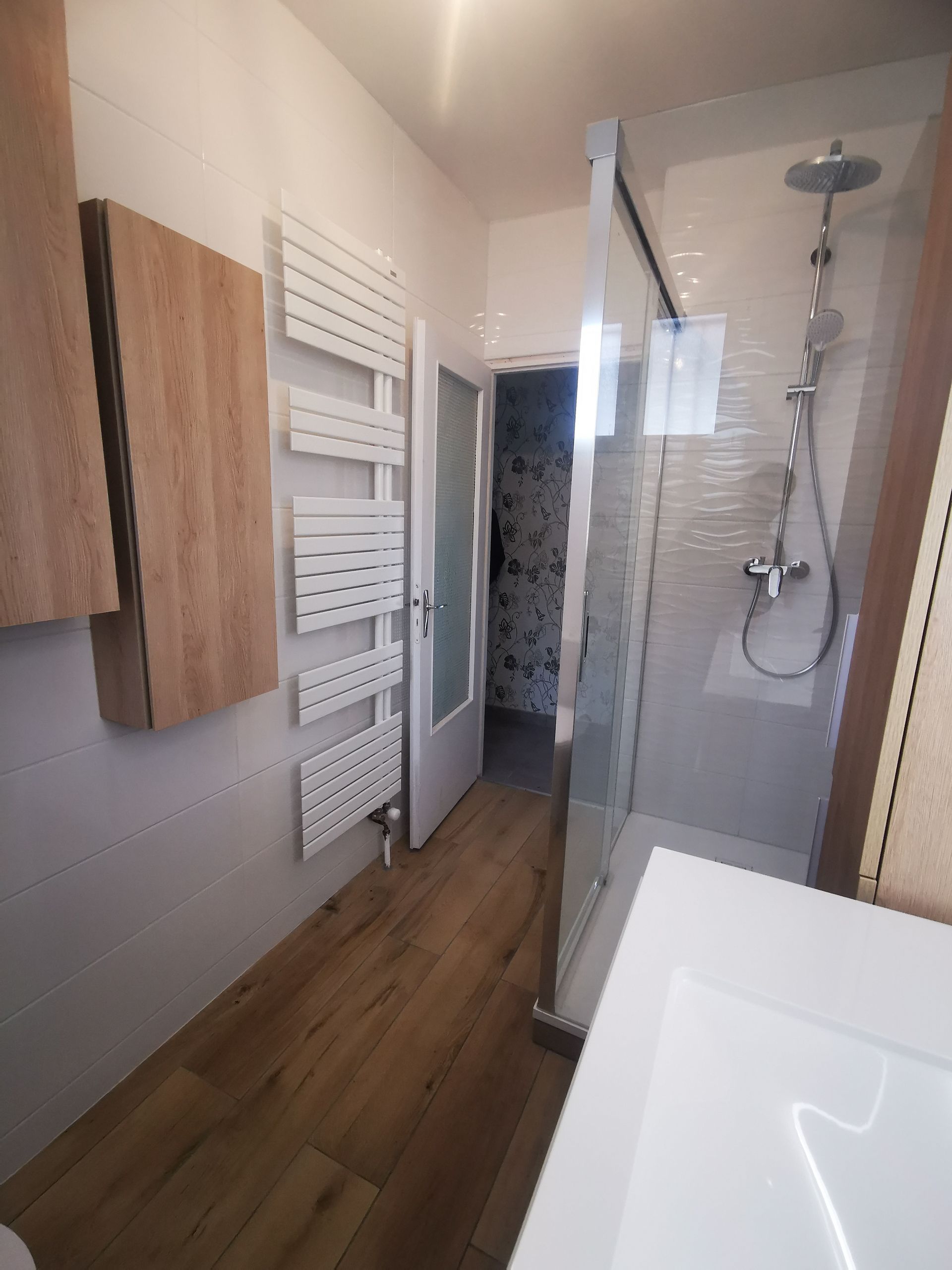 Salle de bain étroite avec douche, radiateur blanc, vanité, plancher imitation bois, armoires en bois clair et porte ouverte.