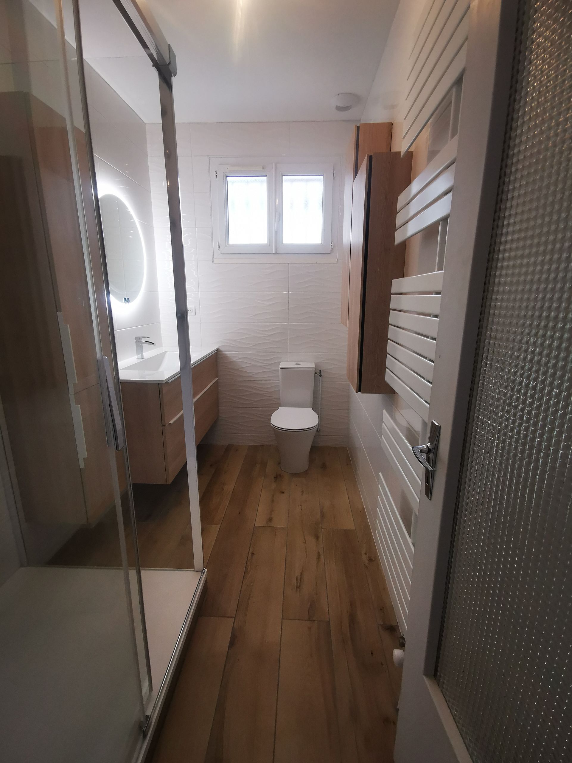 Salle de bain étroite avec sol imitation bois, toilettes blanches, lavabo avec miroir, cabine de douche et sèche-serviettes.