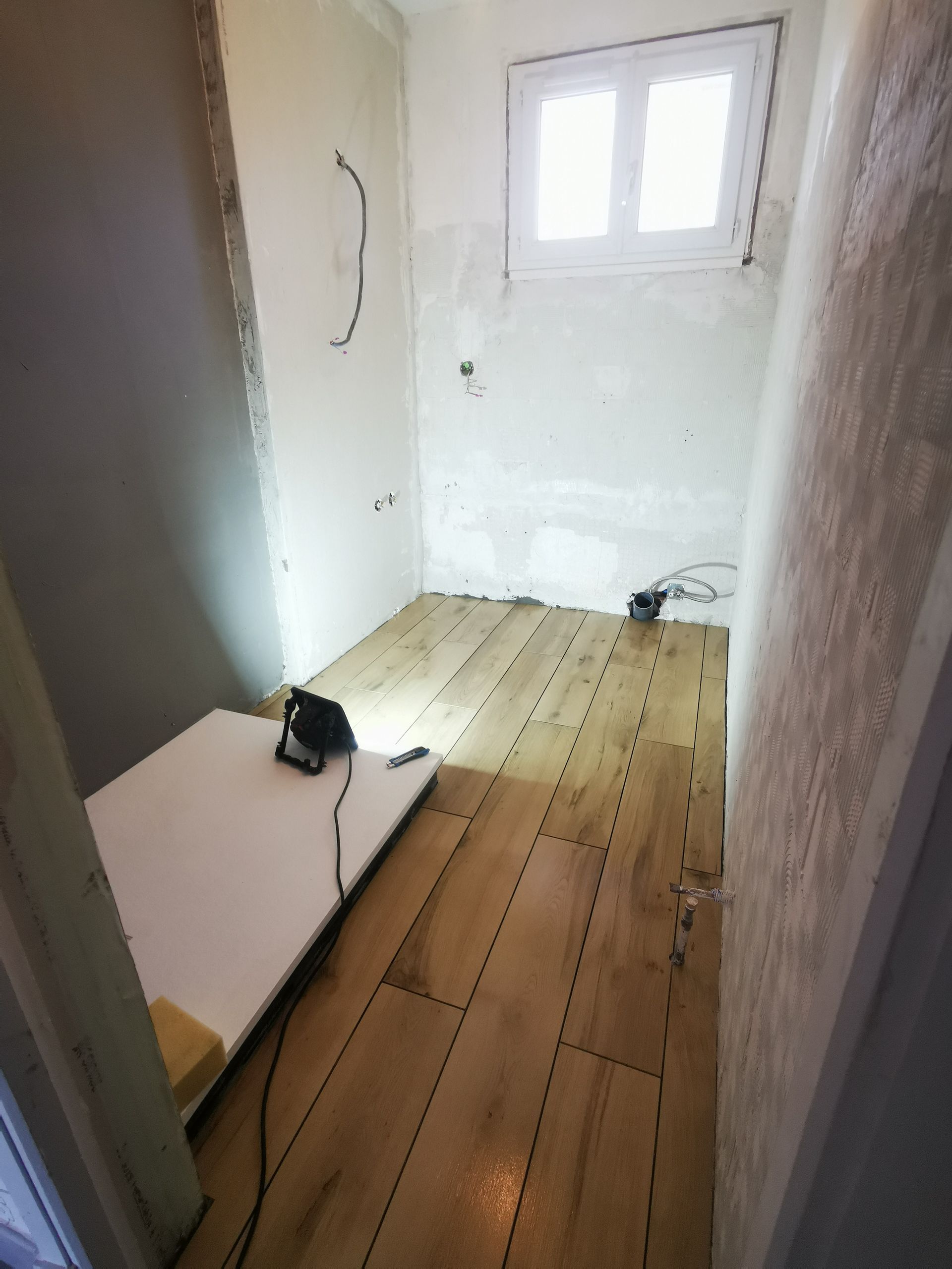 Petite pièce en construction avec parquet imitation bois, murs nus et une petite fenêtre.