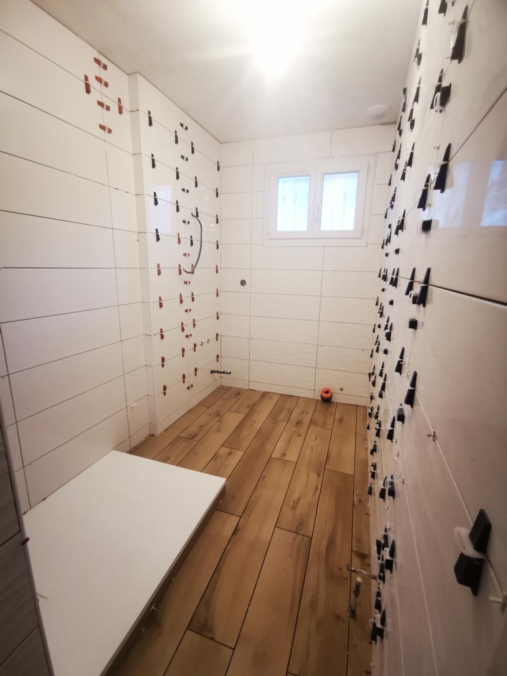 Salle de bain avec murs carrelés blancs, parquet, banc blanc et petite fenêtre. Le carrelage est en cours de pose.