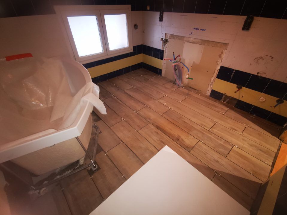 Salle de bains avec carrelage imitation bois et finitions jaune/noir. Une baignoire partiellement installée et une fenêtre sont visibles.