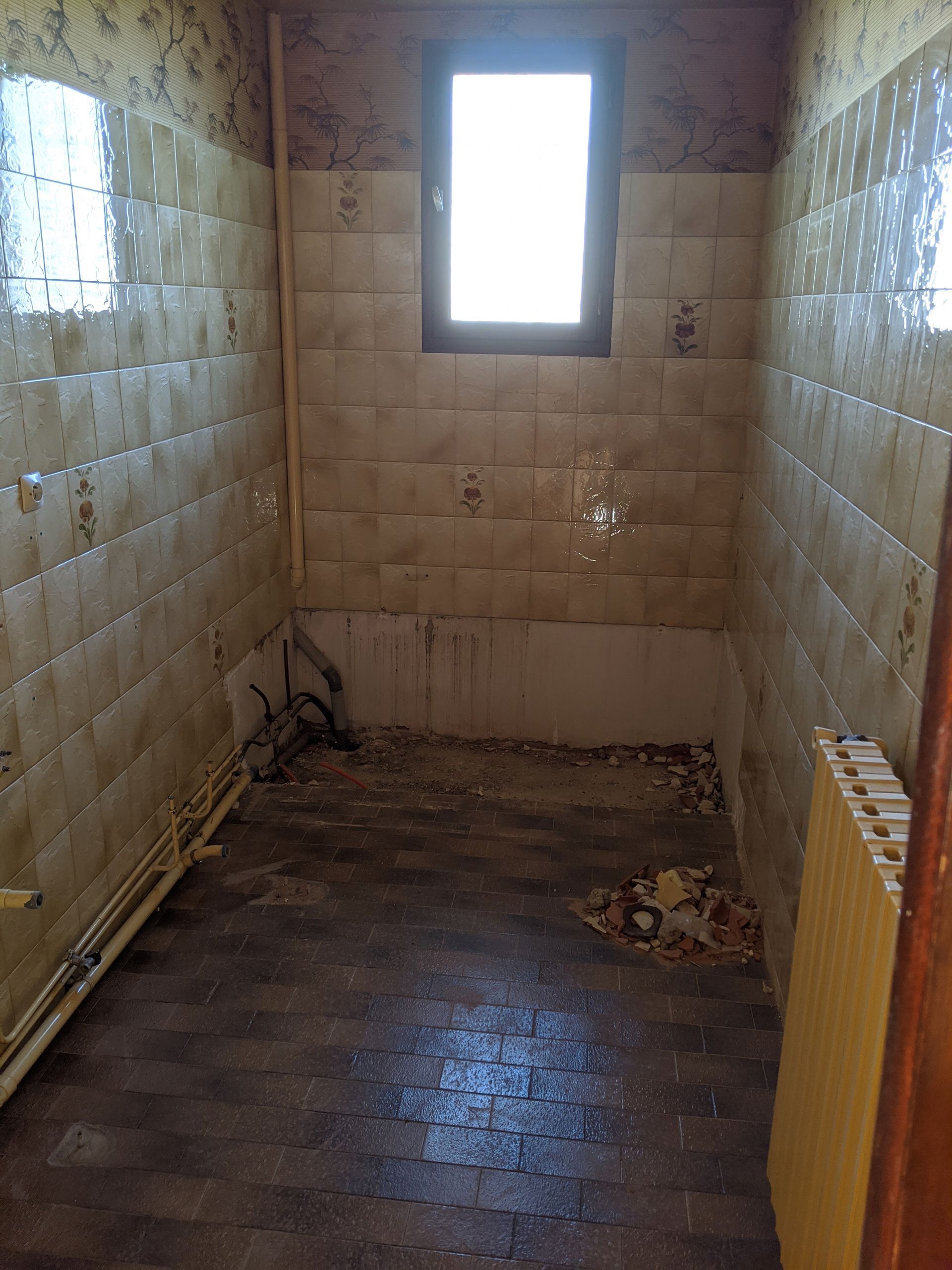 Salle de bain sale et désuète : murs carrelés, petite fenêtre, débris sur le sol, le radiateur et les tuyaux.