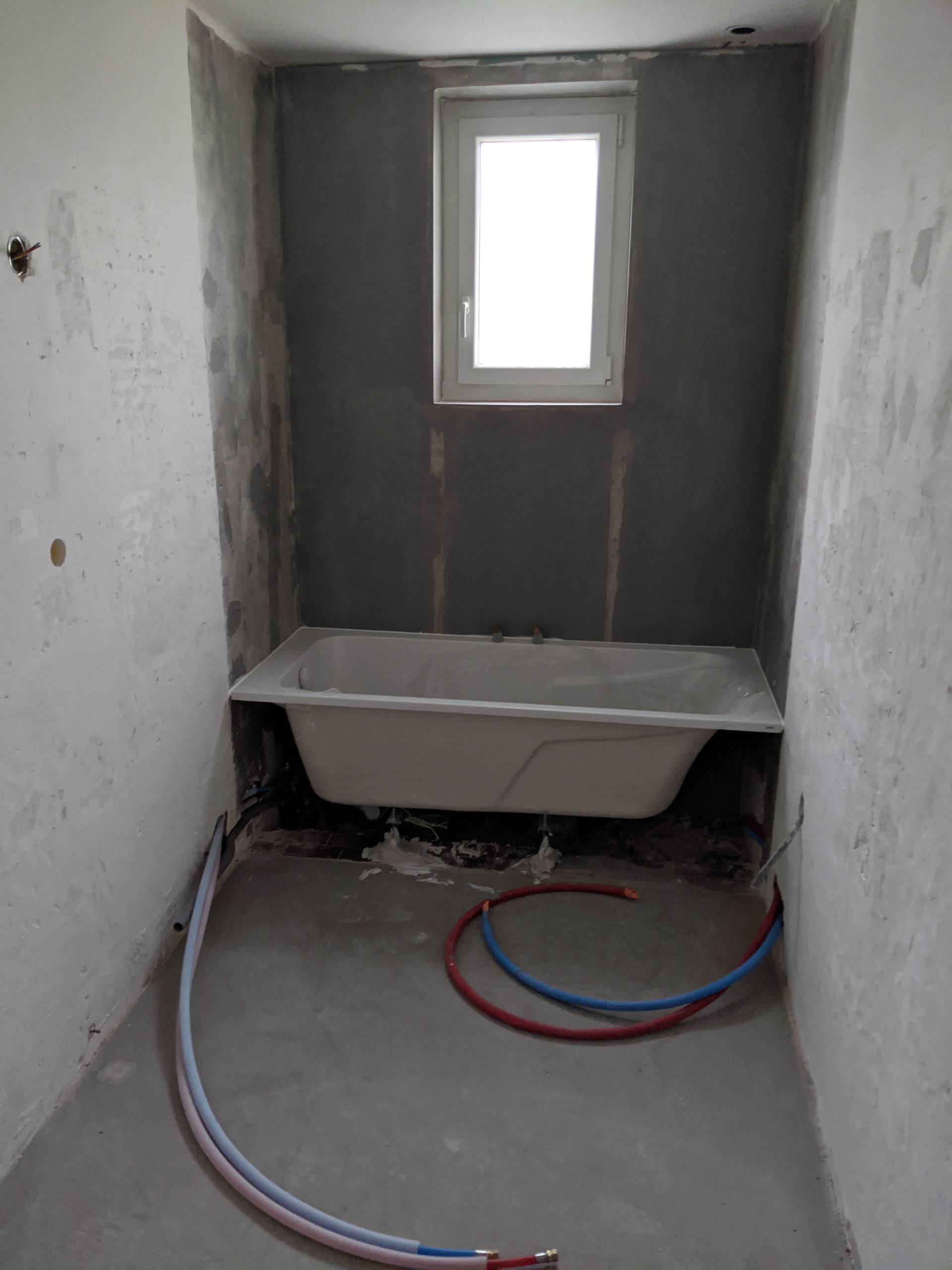 Salle de bain en construction, baignoire sous une fenêtre, plomberie apparente. Murs et sol gris.