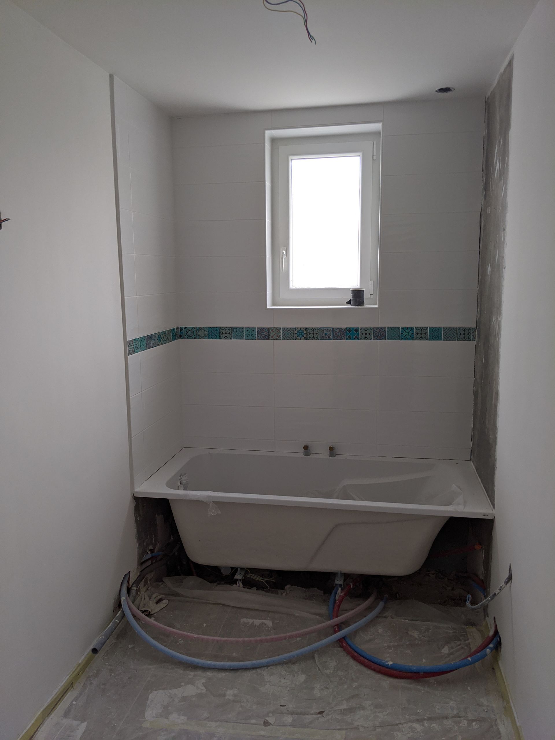 Rénovation de salle de bain en cours : baignoire sous une petite fenêtre, tuyaux bleus et rouges au sol.