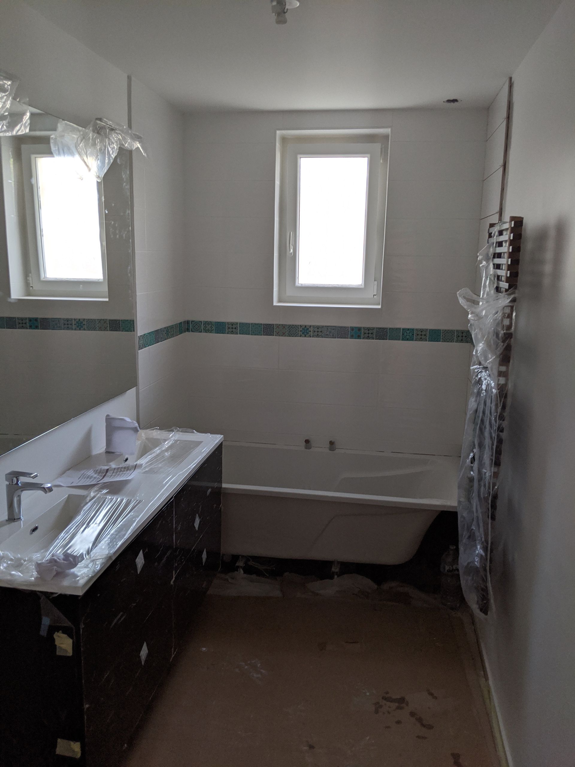 Salle de bain en construction : murs blancs, baignoire, lavabo, fenêtres, bordure de carrelage bleu, sol marron.