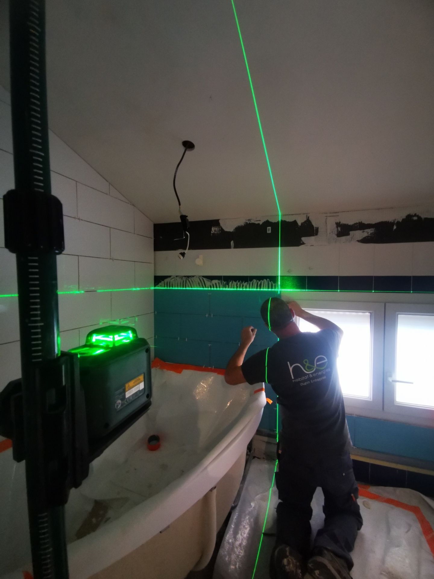 Un homme agenouillé utilise un niveau laser vert pour aligner le carrelage d'une salle de bains. Poutres vert vif, carrelage blanc et bleu sarcelle.
