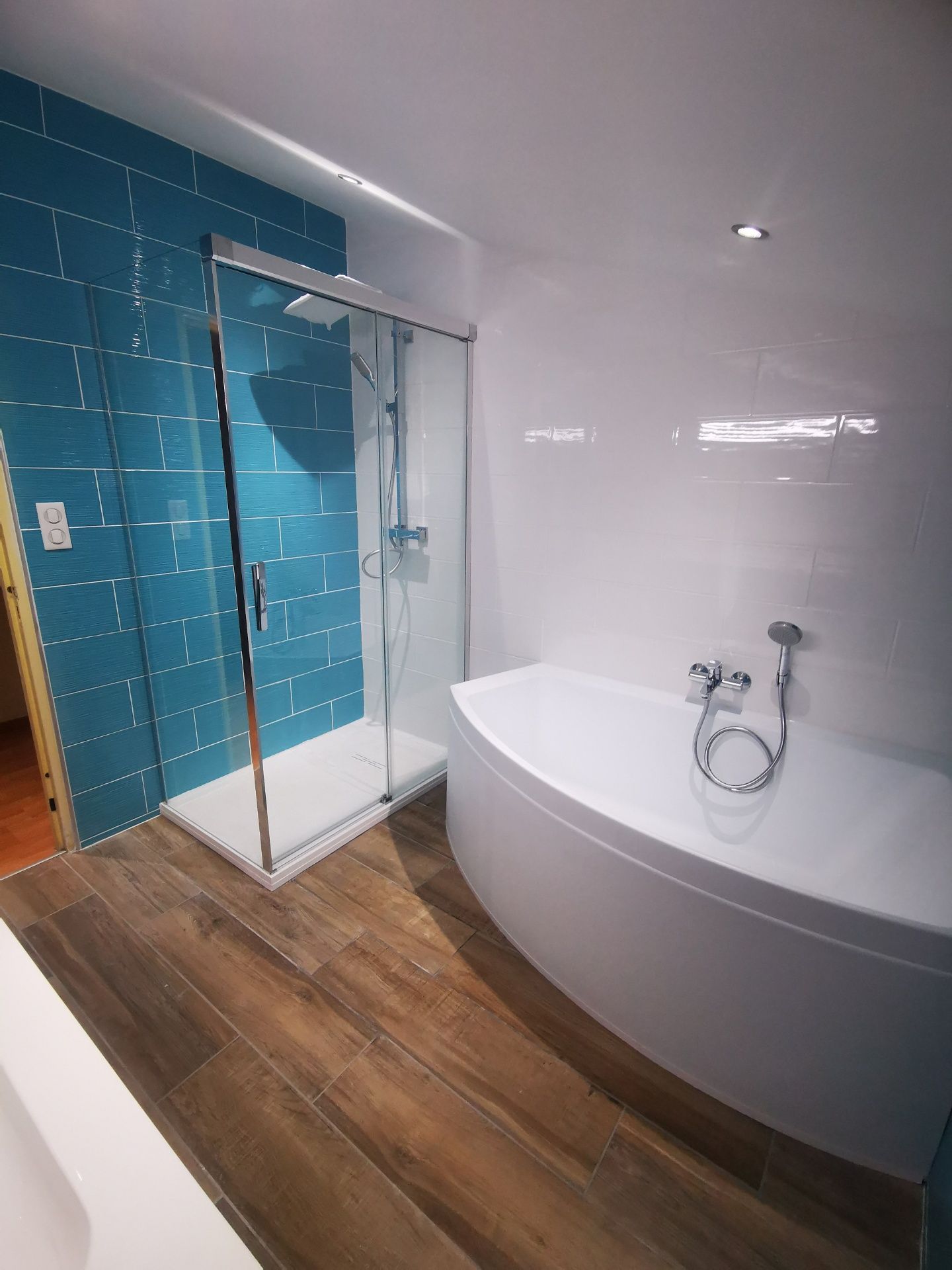Salle de bain moderne avec mur carrelé bleu, douche en verre, baignoire blanche et plancher de bois.