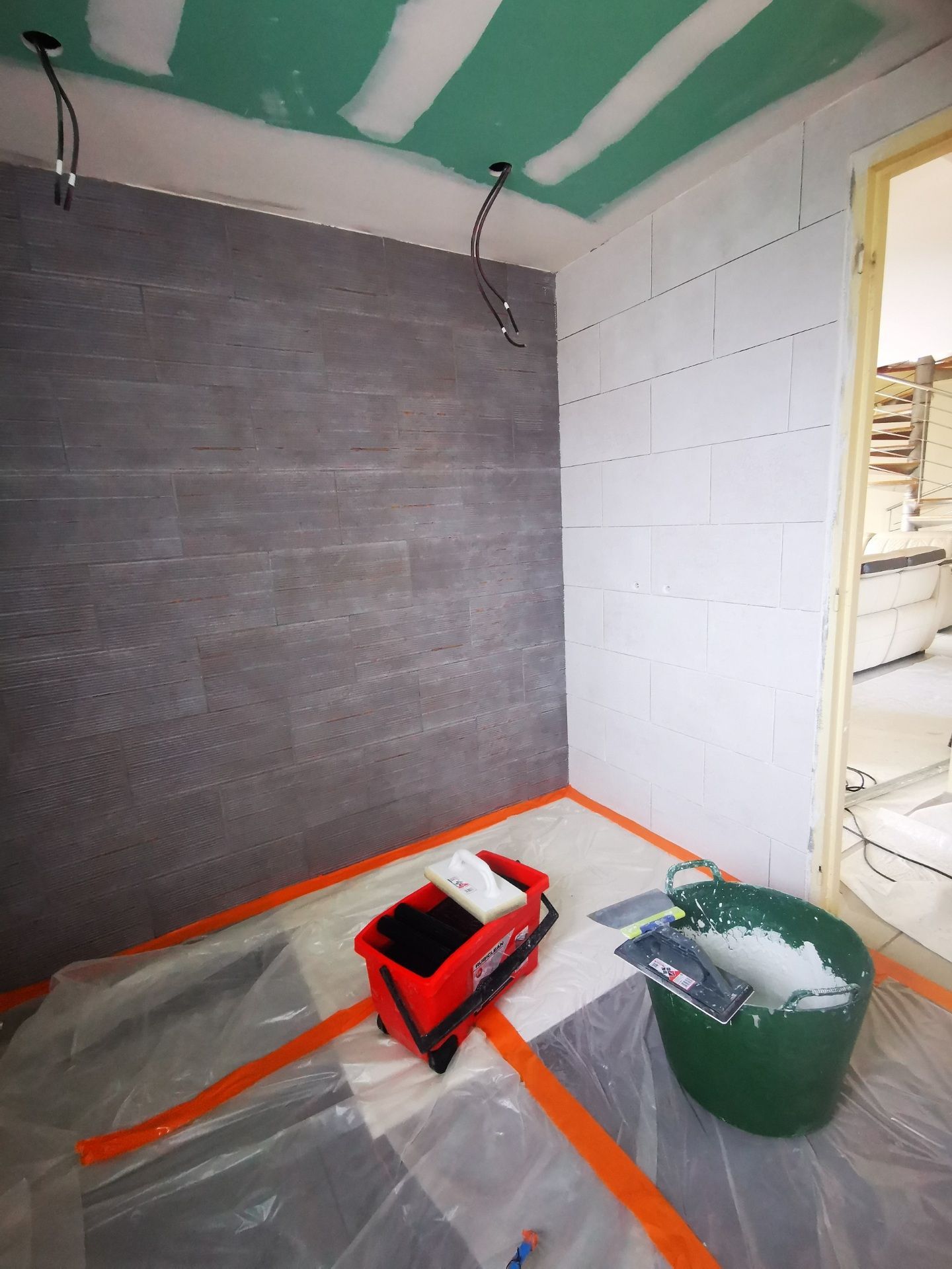 Rénovation de salle de bain en cours, comprenant un mur carrelé gris, des murs en blocs blancs, un plafond vert, du plastique de protection, des outils.
