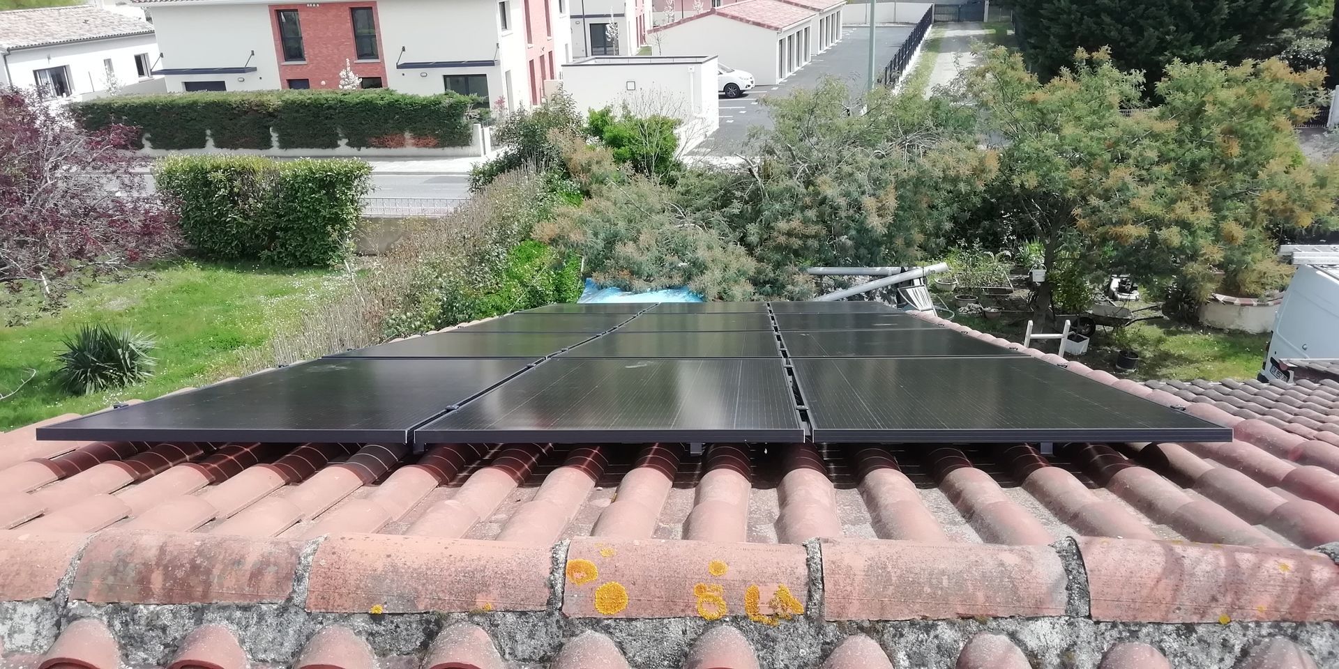 Panneaux solaires installés sur un toit en tuiles rouges, entouré de verdure et de bâtiments en arrière-plan.