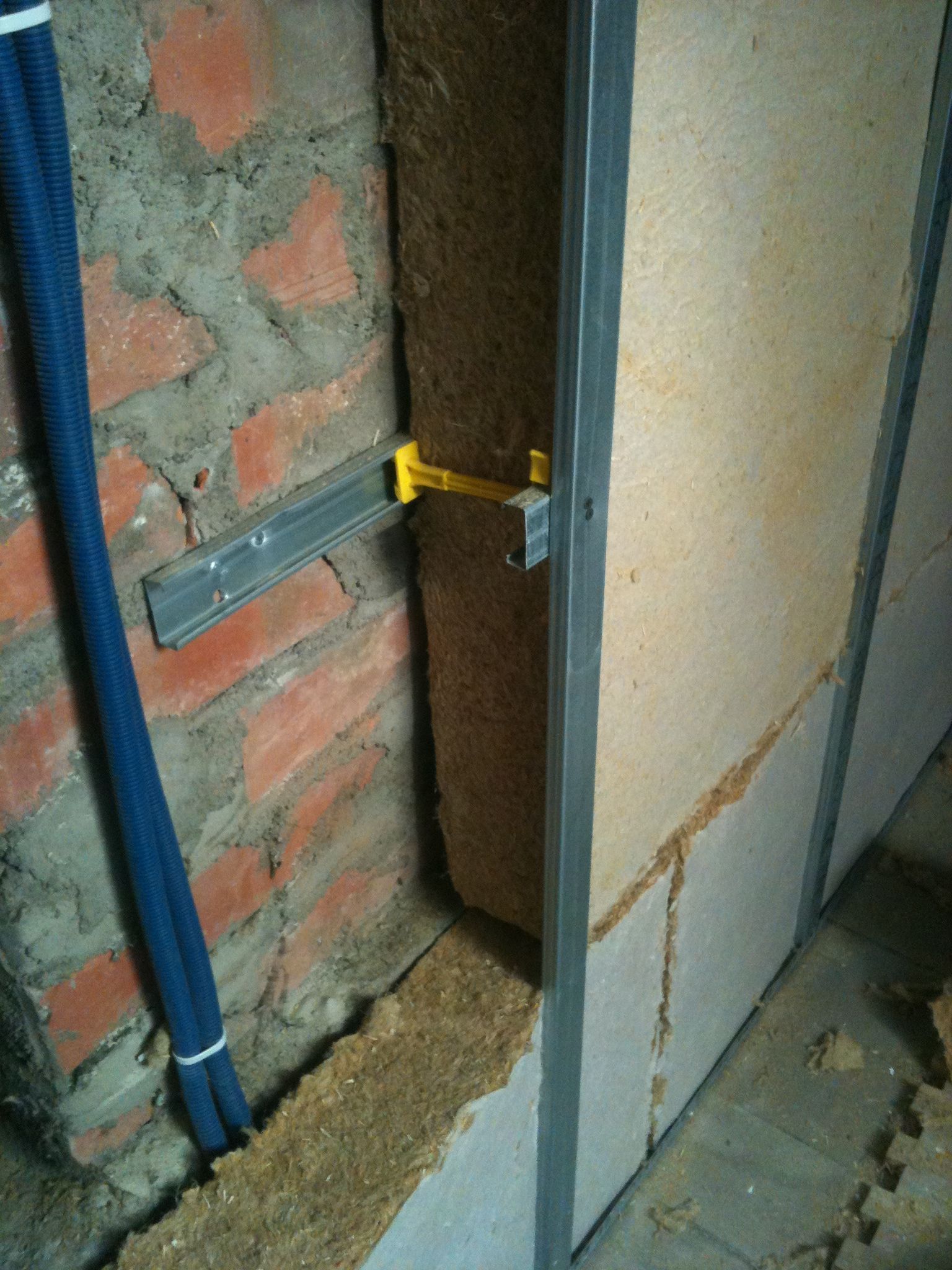 Mur de briques avec isolation et montants métalliques, conduit bleu et ossature en plaques de plâtre en cours.