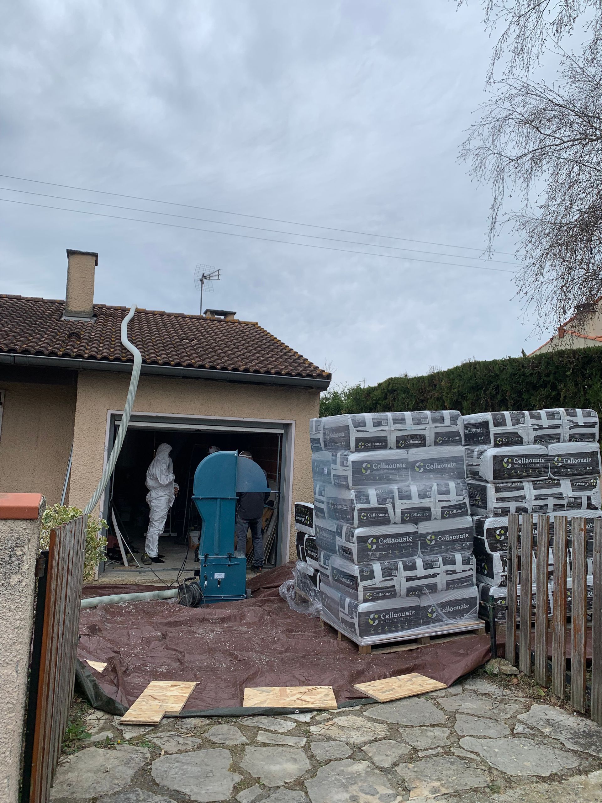 Chantier : ouvriers en combinaison de protection dans un garage, briques empilées à l'extérieur. Ciel couvert.