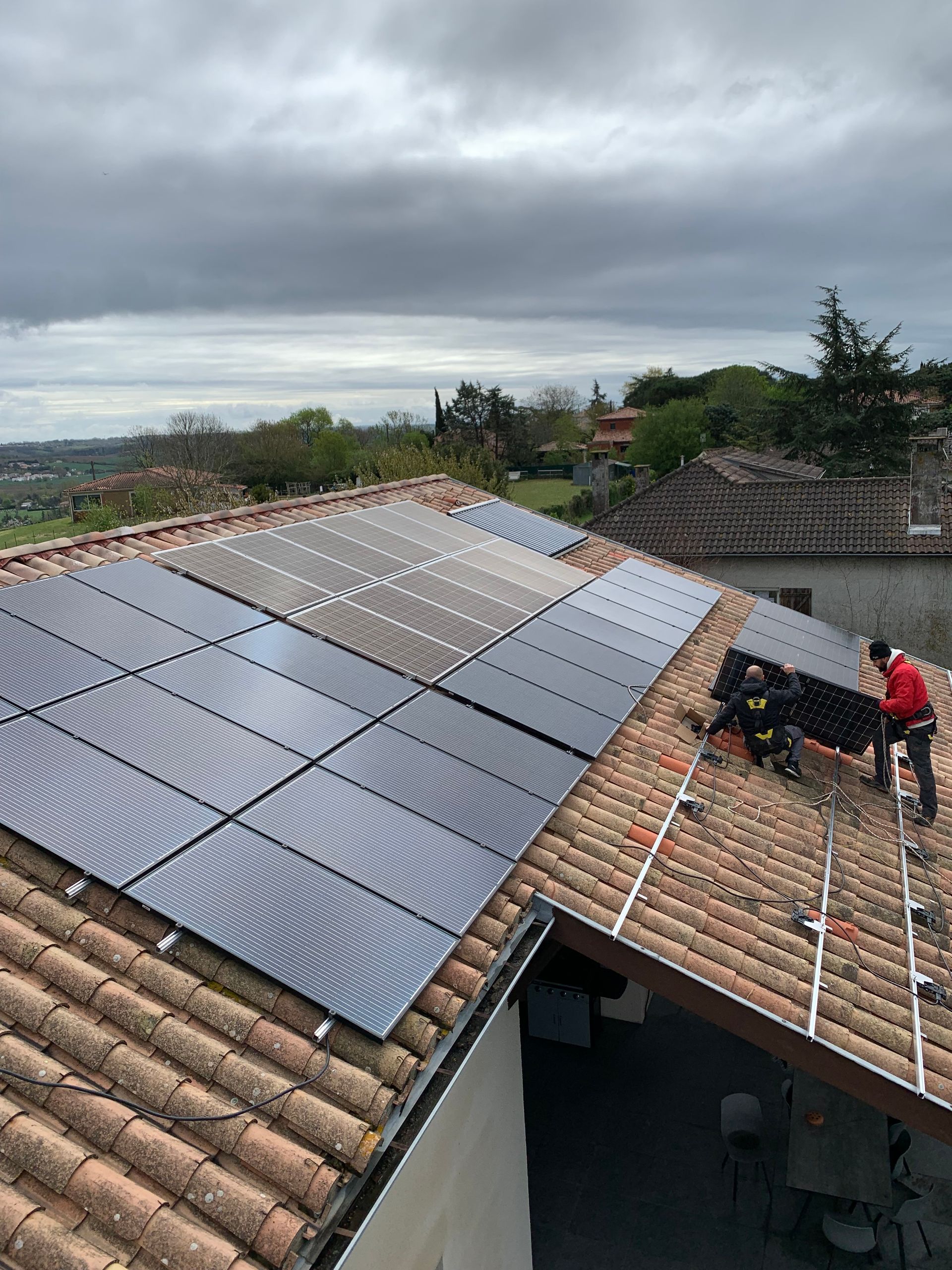 Des ouvriers installent des panneaux solaires sur un toit en tuiles sous un ciel nuageux.