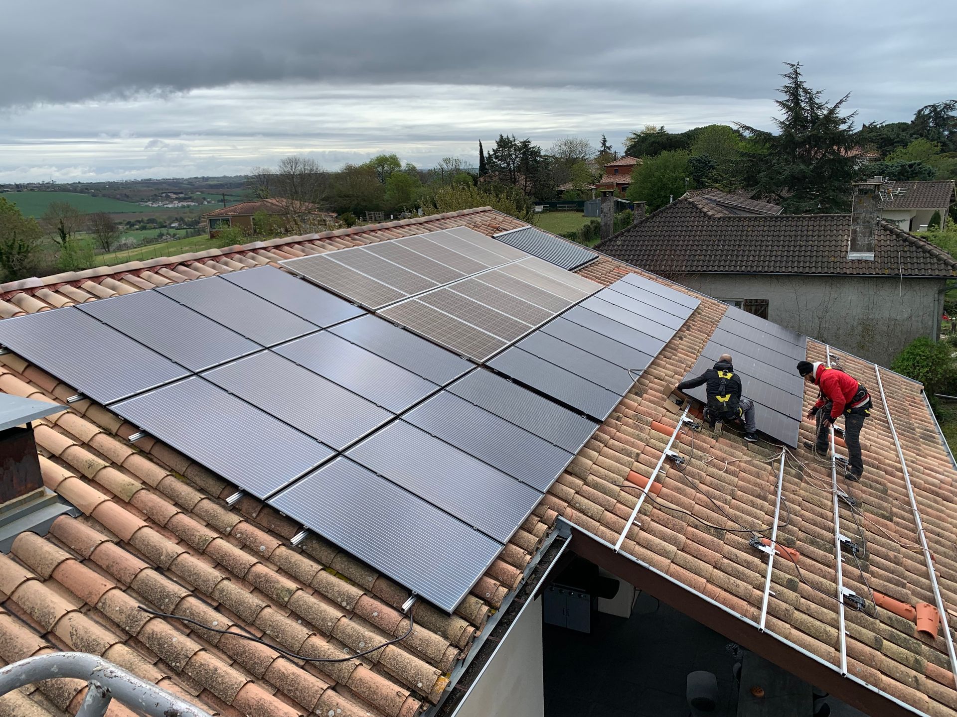Installation de panneaux solaires sur un toit en tuiles de terre cuite par deux ouvriers en tenue de sécurité. Ciel nuageux en arrière-plan.