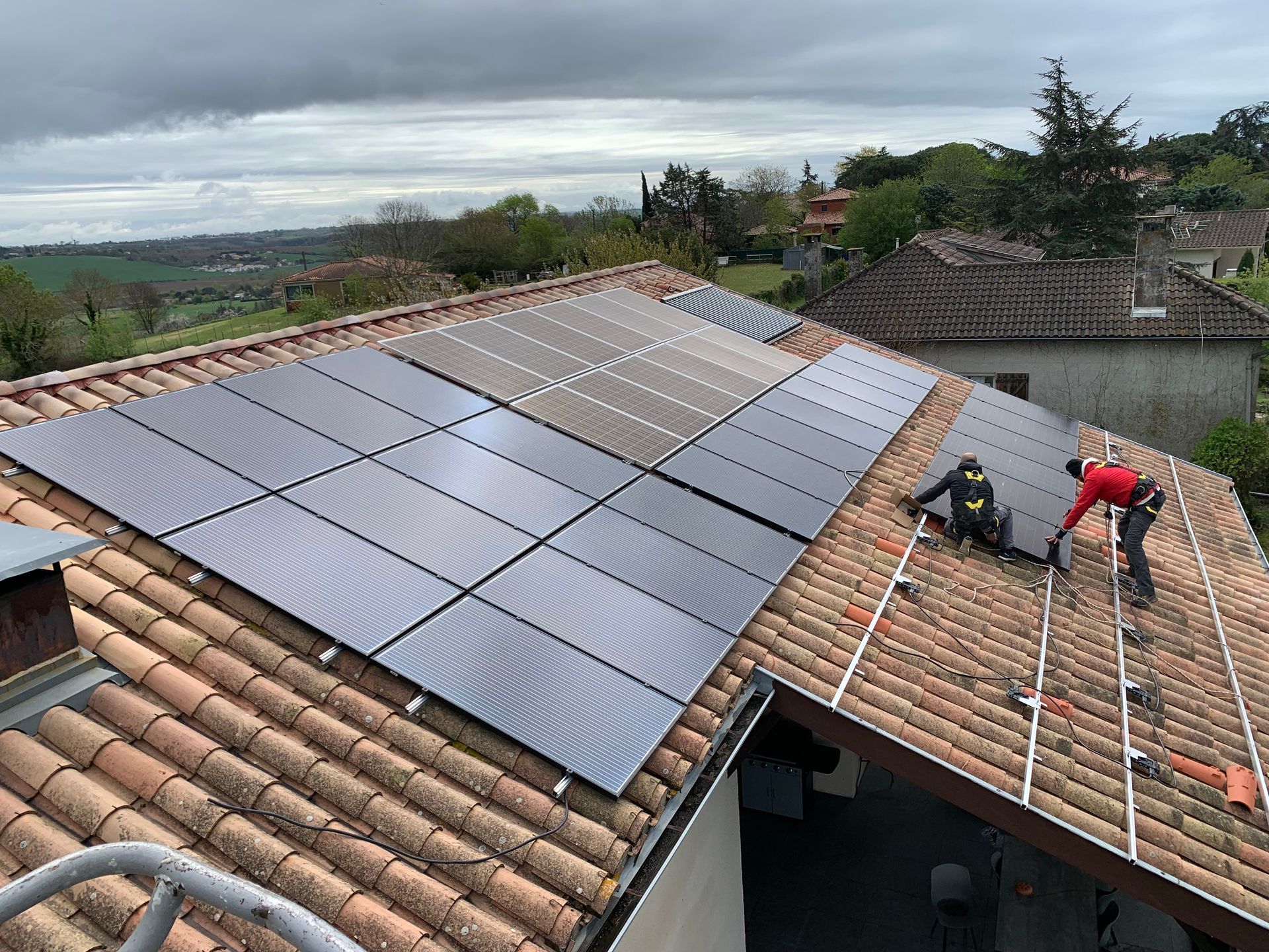 Des panneaux solaires sont installés sur un toit en tuiles en terre cuite par deux ouvriers par une journée nuageuse.