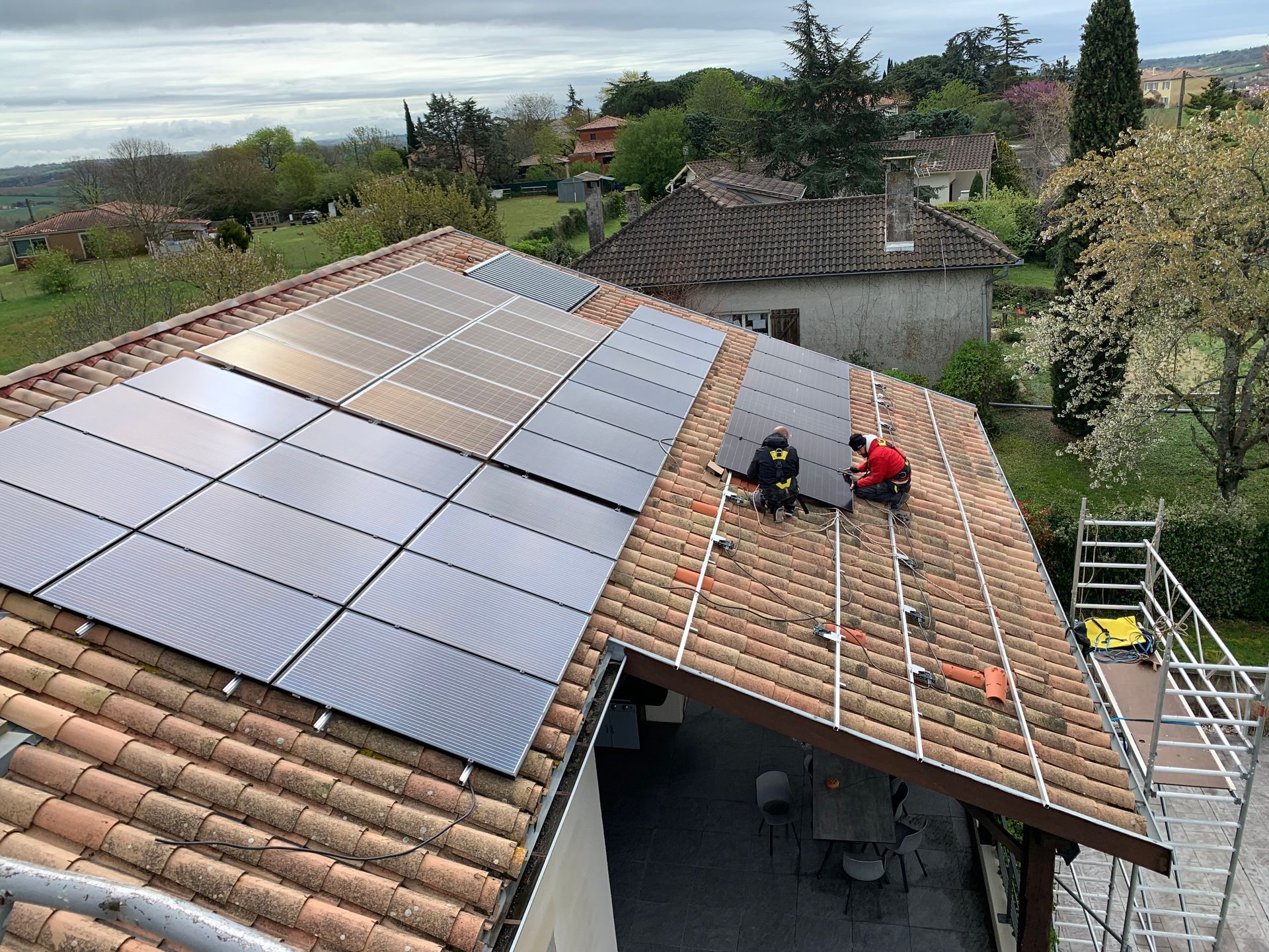 Des ouvriers installent des panneaux solaires sur un toit en tuiles en terre cuite, avec des maisons et des arbres visibles en arrière-plan.