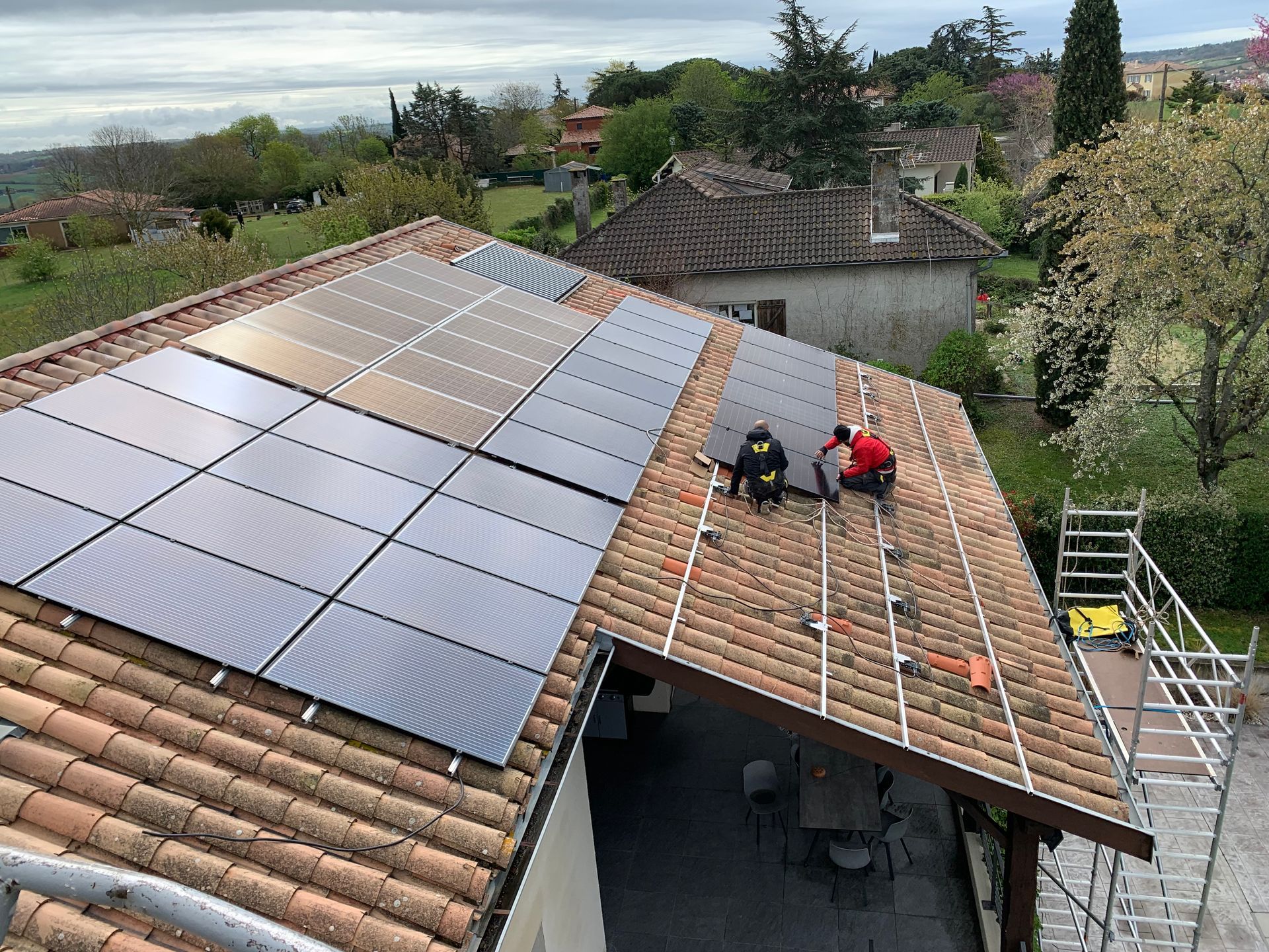 Installation de panneaux solaires sur un toit en tuiles par deux ouvriers. Ciel couvert, maisons et arbres en arrière-plan.