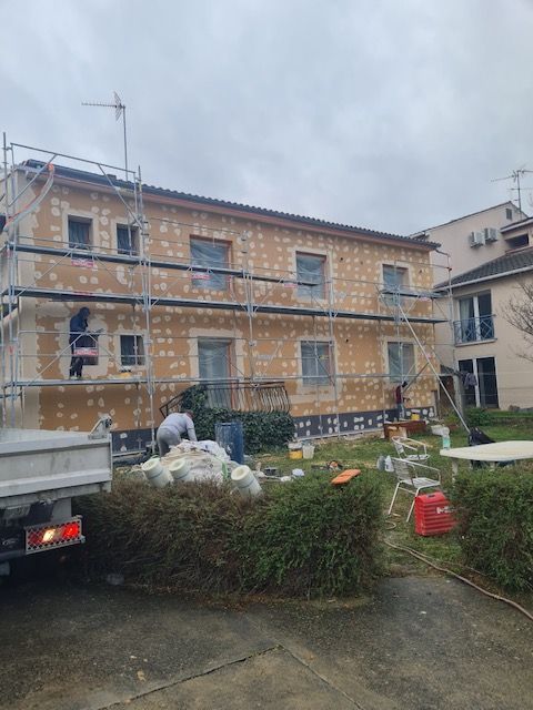 Façade d'immeuble en rénovation, avec échafaudages, ouvriers et matériaux. Ciel nuageux, bâtiment beige, verdure.