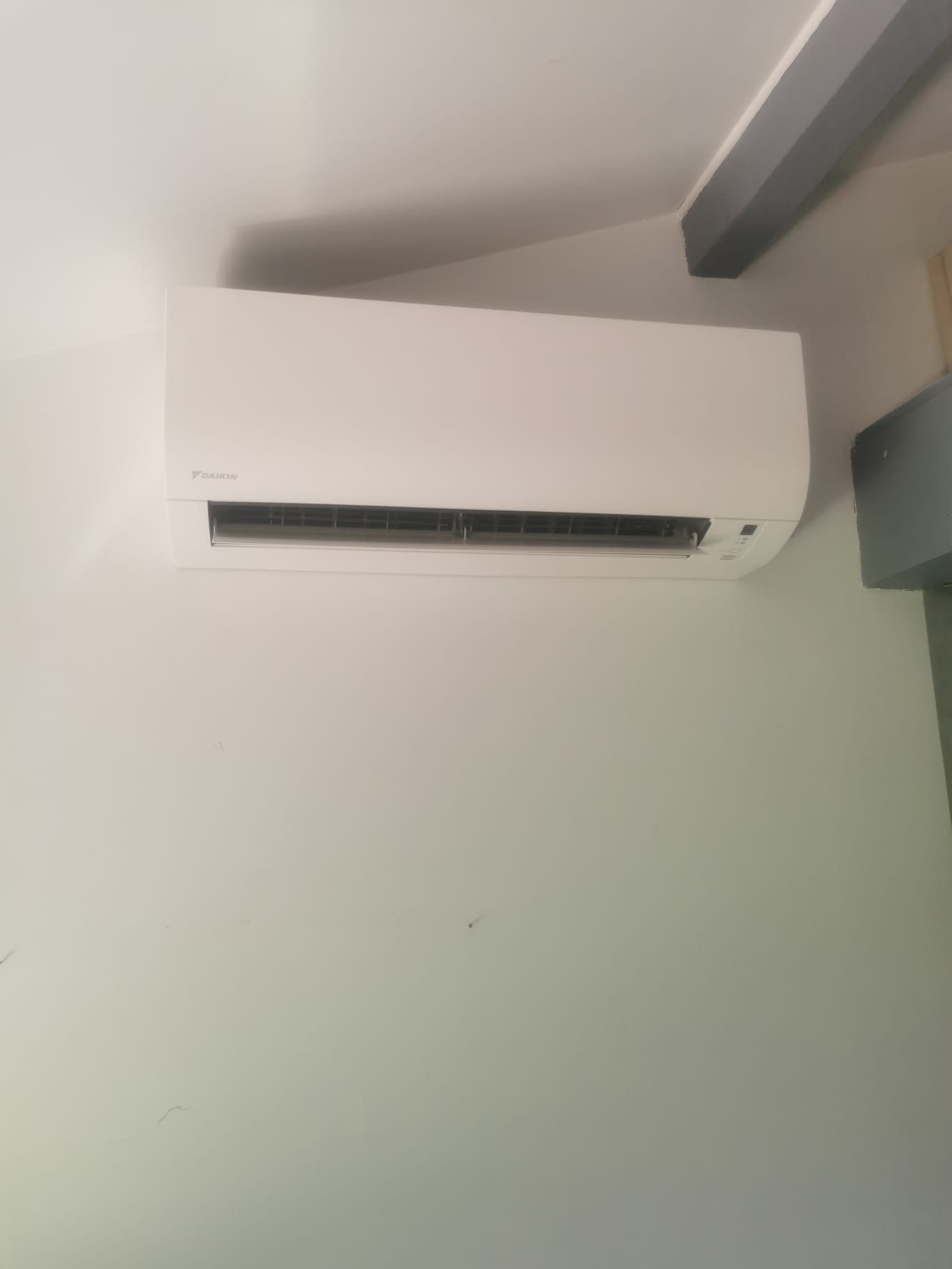 Climatiseur mural blanc sur un mur blanc, avec une poutre de support grise au-dessus.