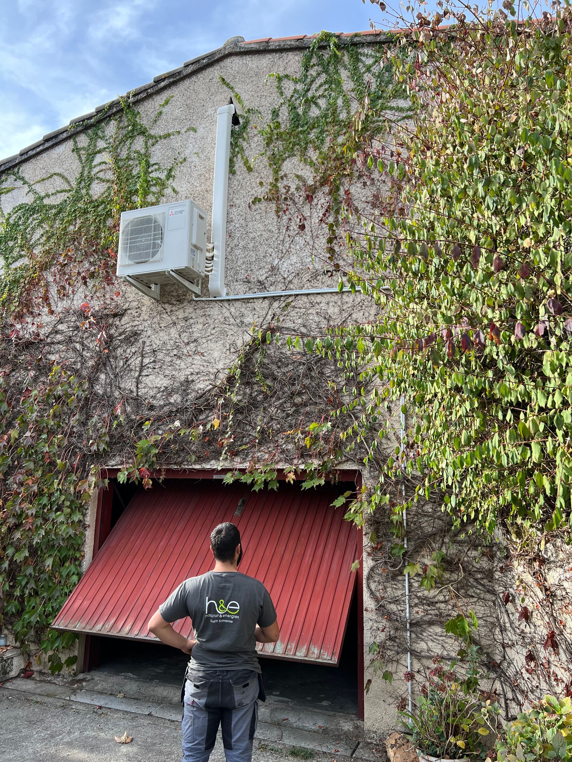 Un homme regarde une porte de garage rouge ouverte sur un bâtiment couvert de vigne avec une unité de climatisation.