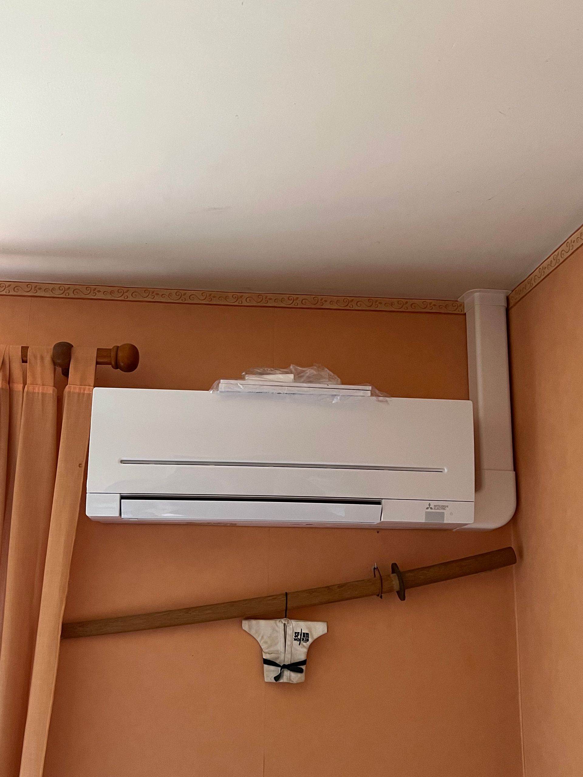 Climatiseur blanc monté sur un mur orange avec une épée katana en dessous.