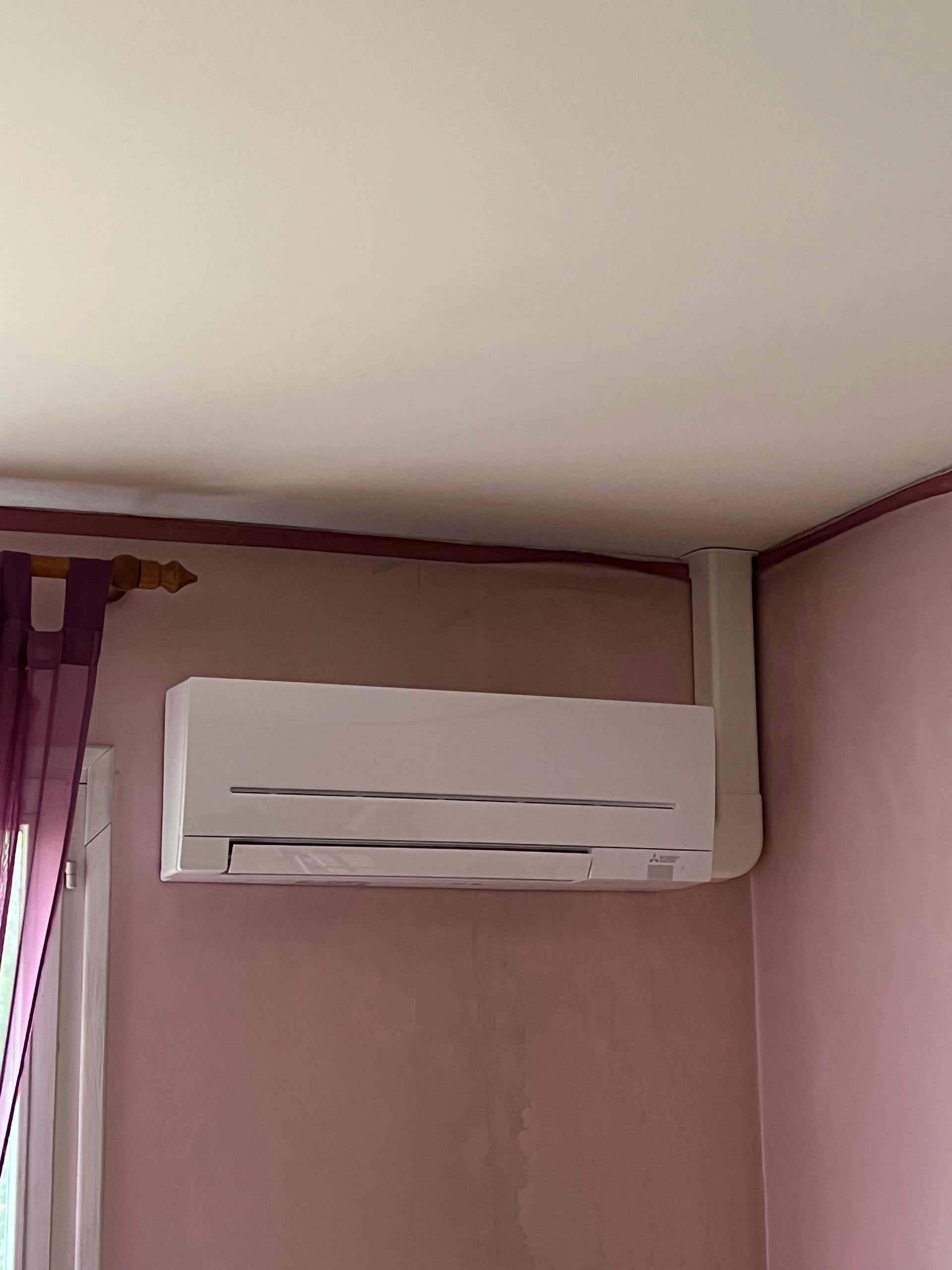 Climatiseur blanc sans conduit monté sur un mur rose, avec un rideau violet à gauche.