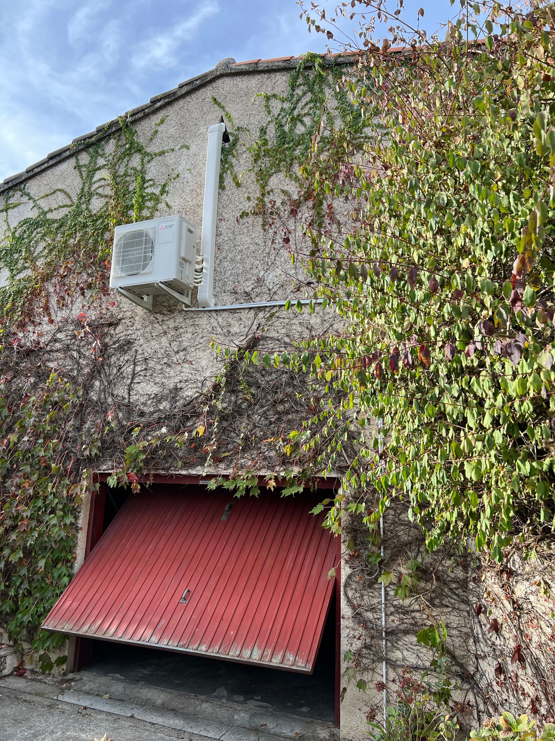 Porte de garage rouge entrouverte dans un bâtiment en stuc recouvert de vigne vierge. Climatisation blanche installée au-dessus.