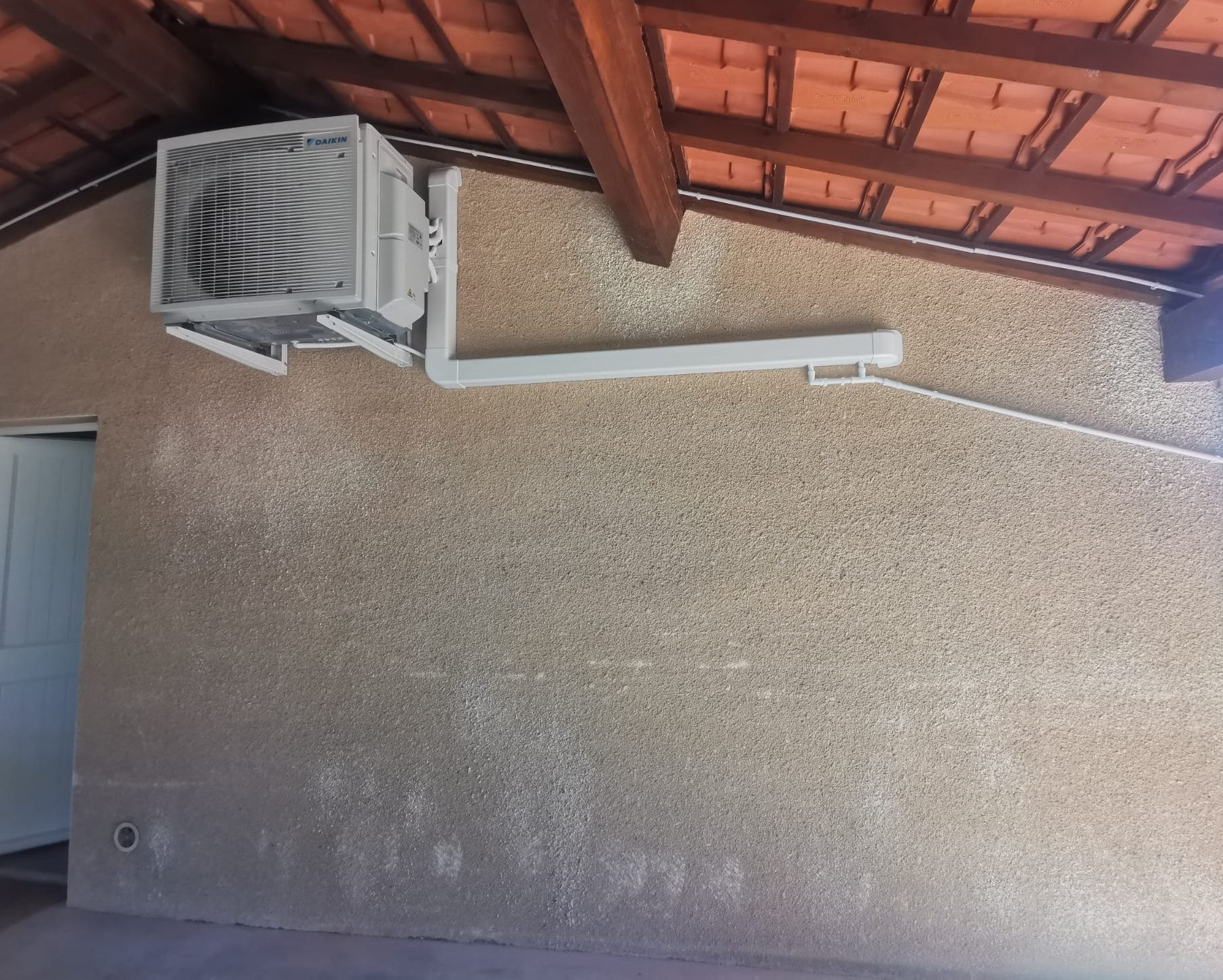 Un refroidisseur d'air monté sur un mur de couleur claire, sous un toit en tuiles.