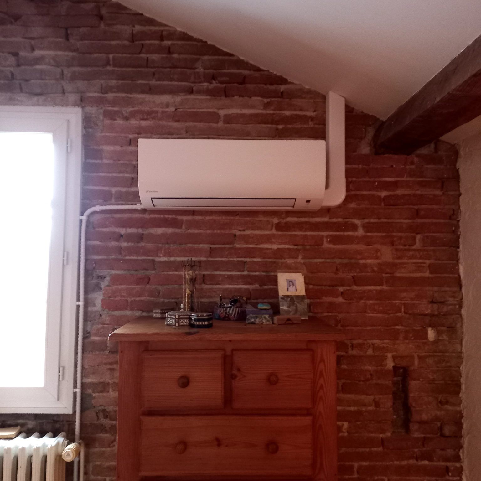 Climatiseur blanc sur un mur de briques rouges au-dessus d'une commode en bois, à côté d'une fenêtre.