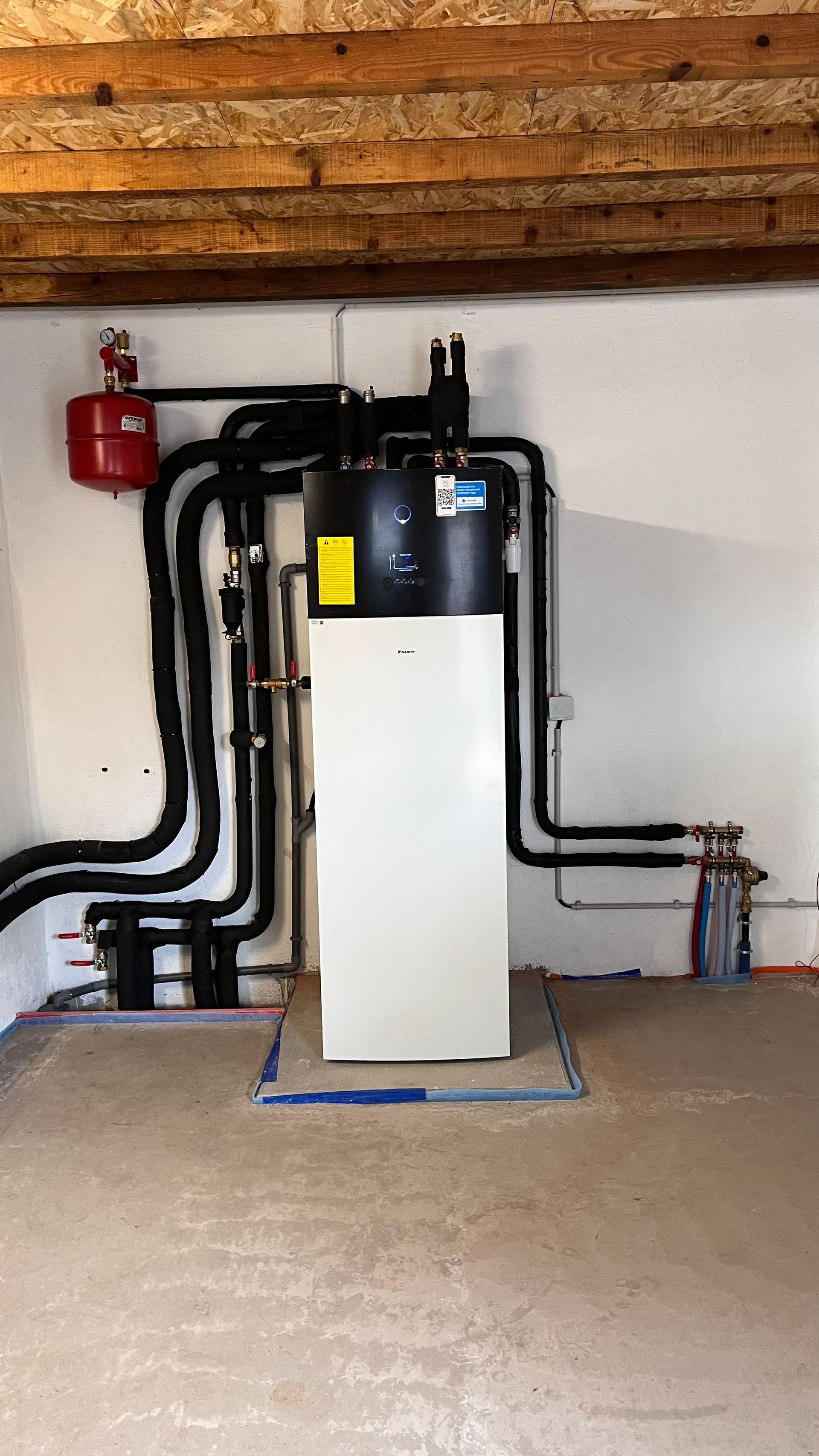 Système de pompe à chaleur blanc et noir avec vase d'expansion rouge contre un mur blanc dans une pièce avec poutres en bois.