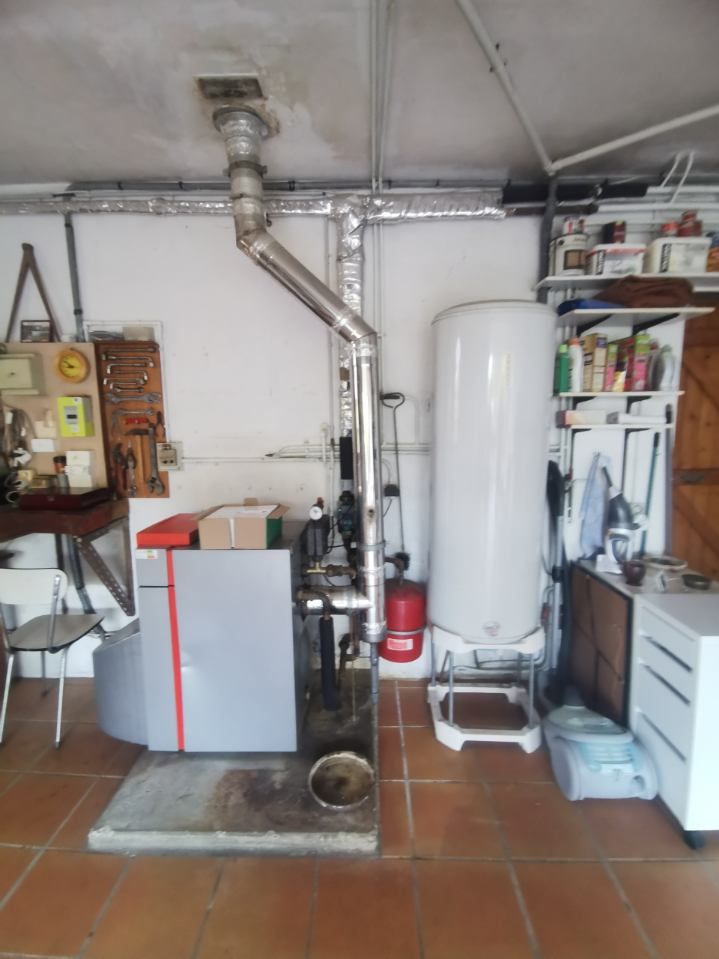 Fournaise et chauffe-eau dans un garage. Électroménagers gris et blancs, cheminée en inox, outils accrochés au mur.