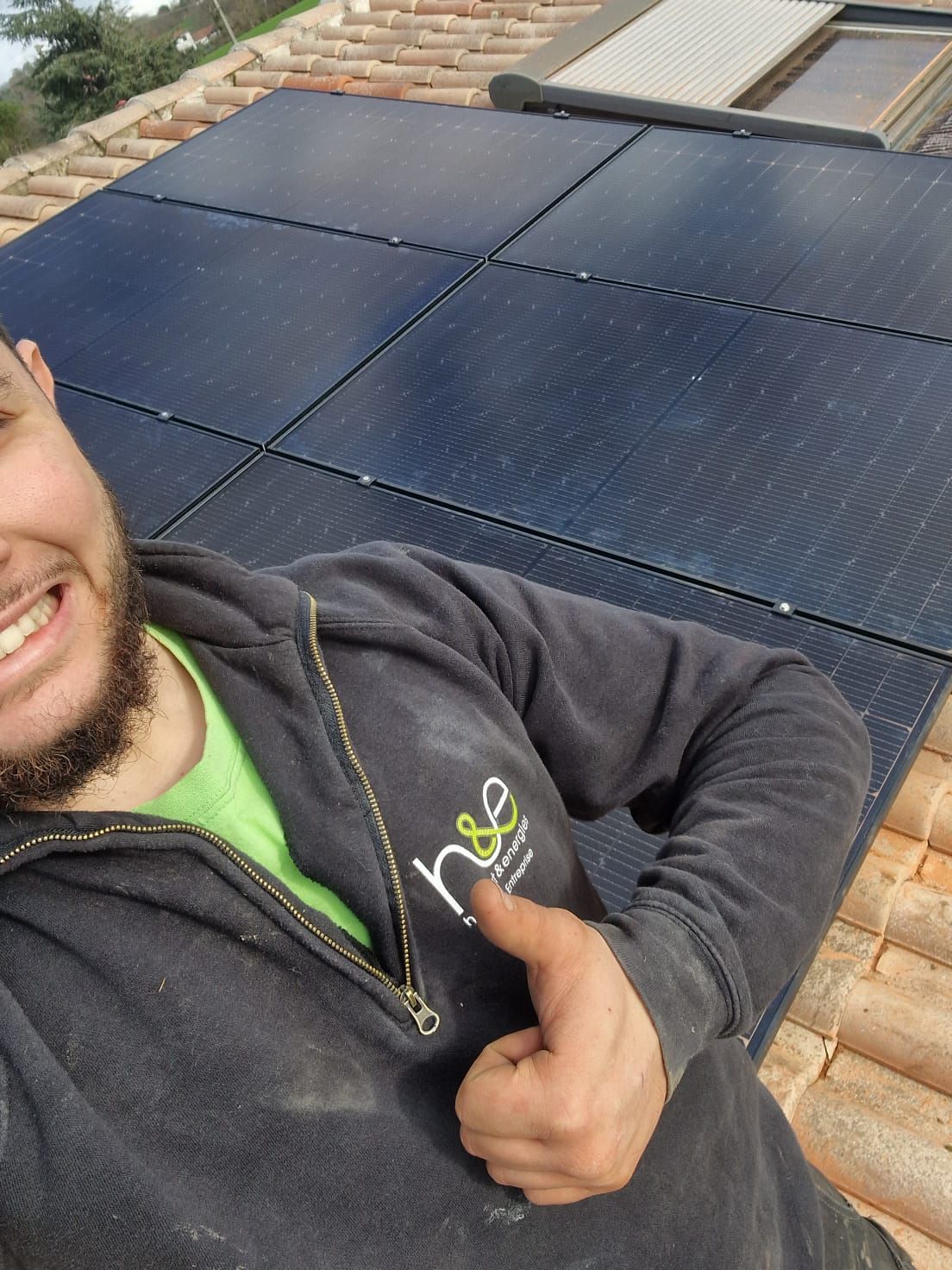 Homme sur un toit avec des panneaux solaires, pouce levé. Vêtu d'une veste noire et d'une chemise verte, il sourit.