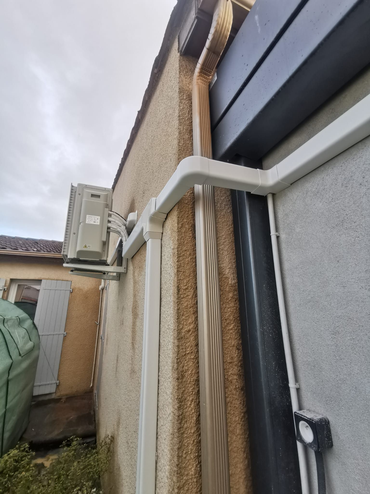 Mur extérieur avec garniture blanche, tuyau de descente marron et unité de climatisation.