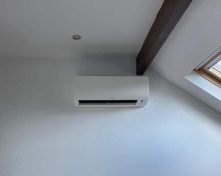 Climatiseur blanc monté sur un mur gris pâle, près d'une poutre en bois foncé et d'un puits de lumière.