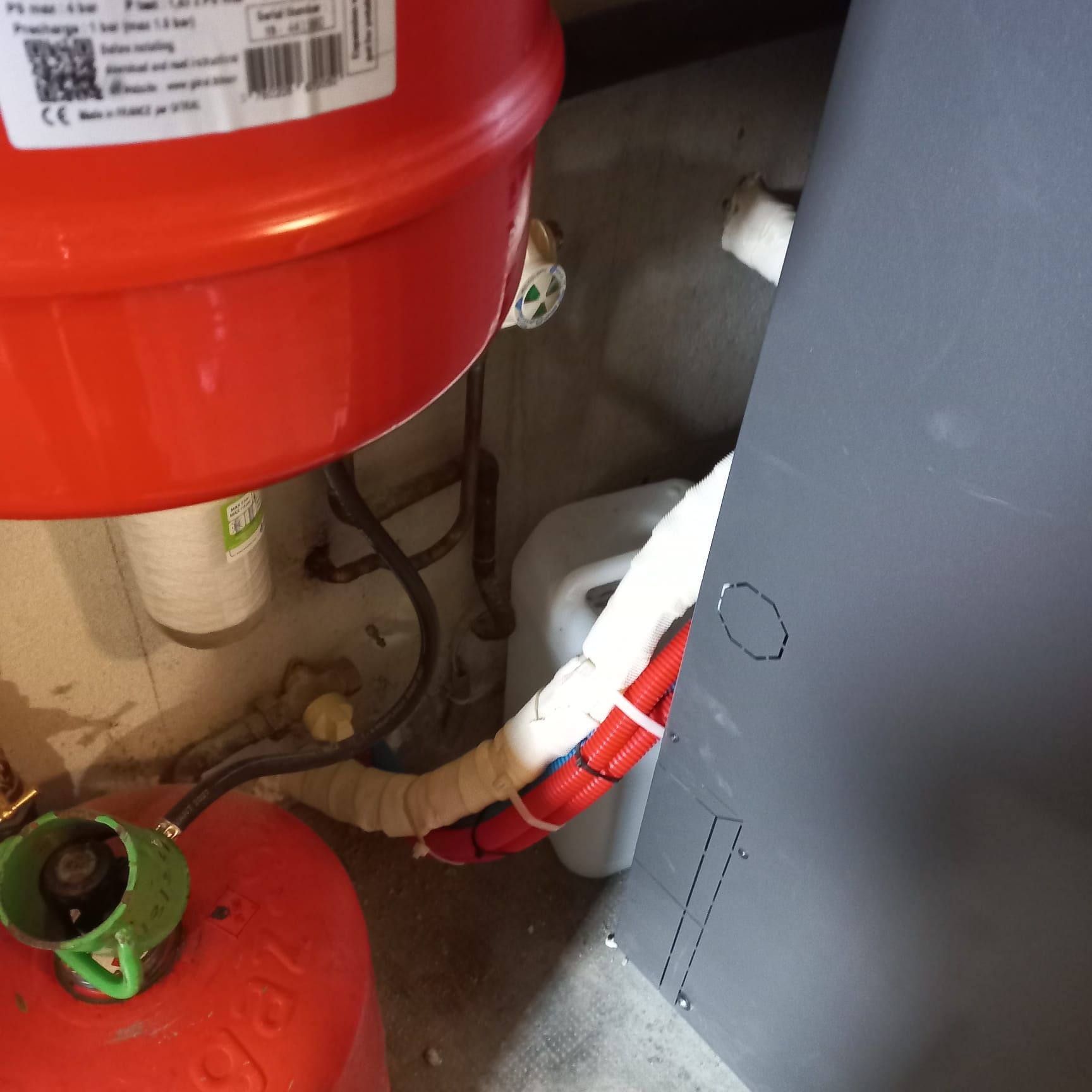 Réservoirs rouges et verts avec tuyaux et un panneau gris, faisant probablement partie d'un système de chauffage dans un sous-sol.
