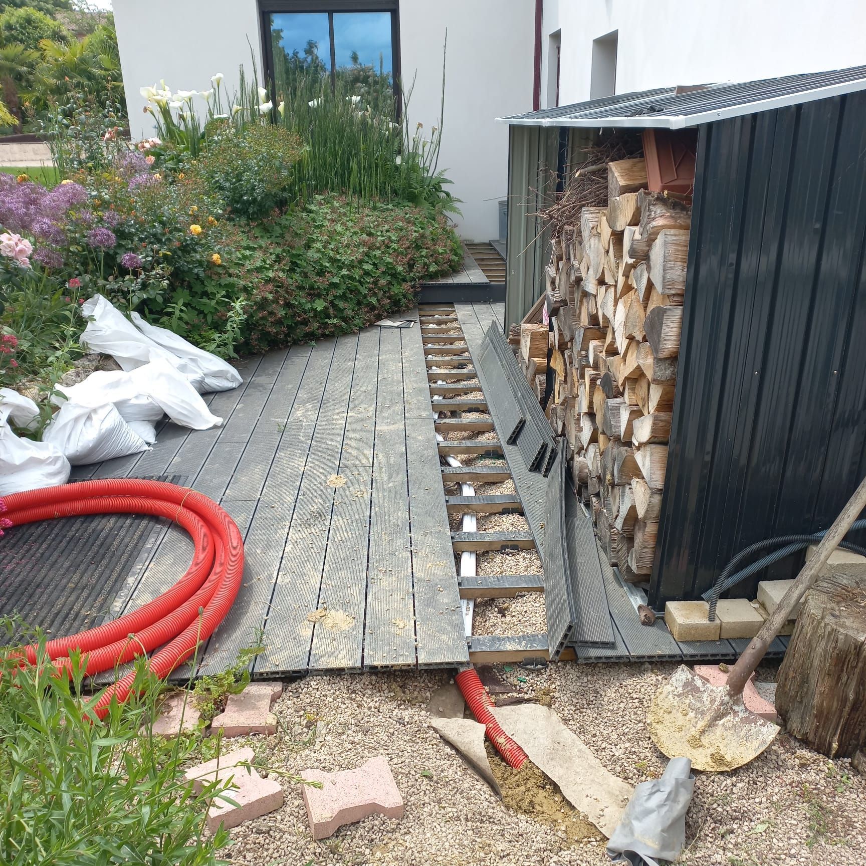 Une terrasse en bois en construction, avec un abri à bois à droite, des tuyaux et des sacs rouges.