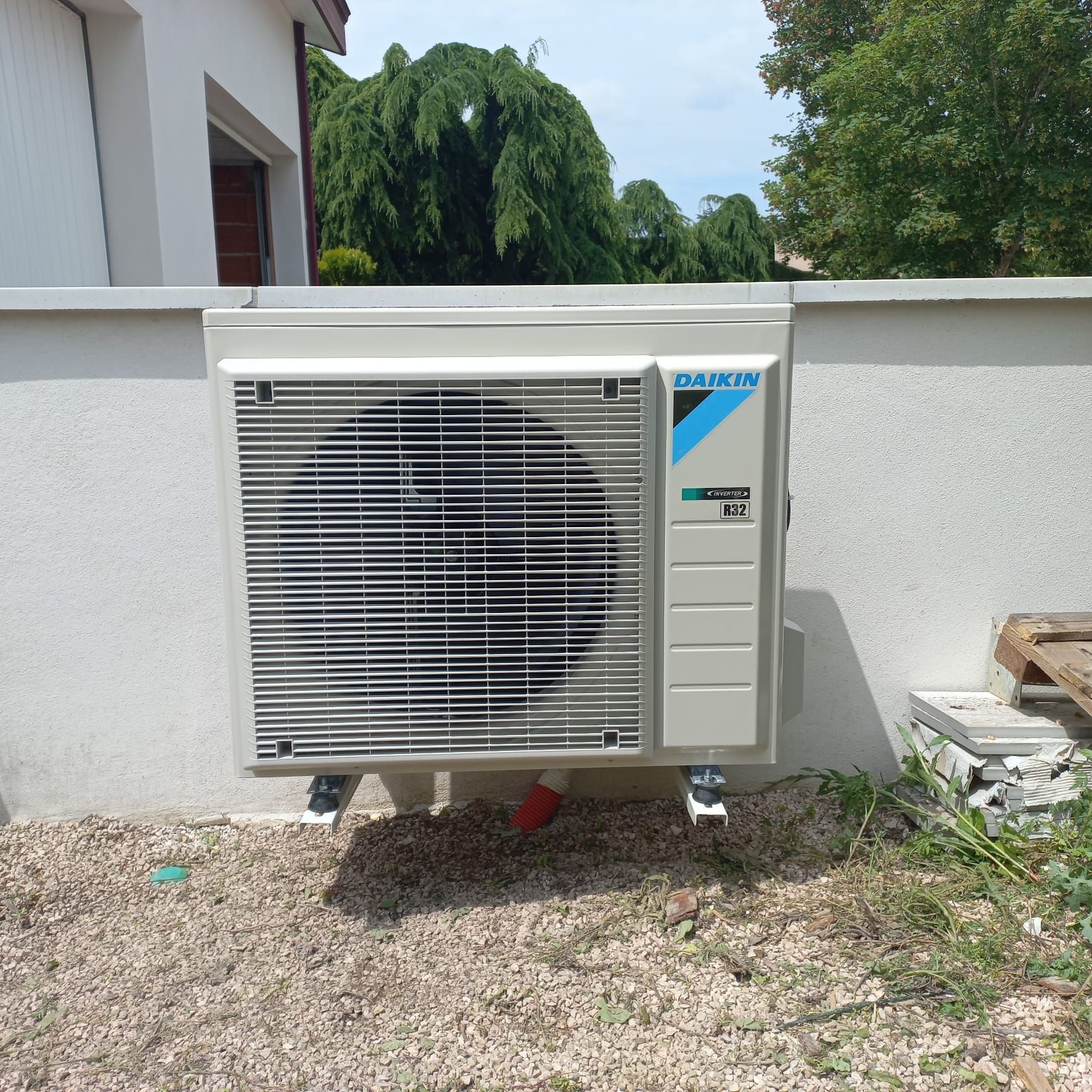 Un climatiseur extérieur Daikin fixé sur un mur blanc. Il est entouré de gravier et d'herbe.