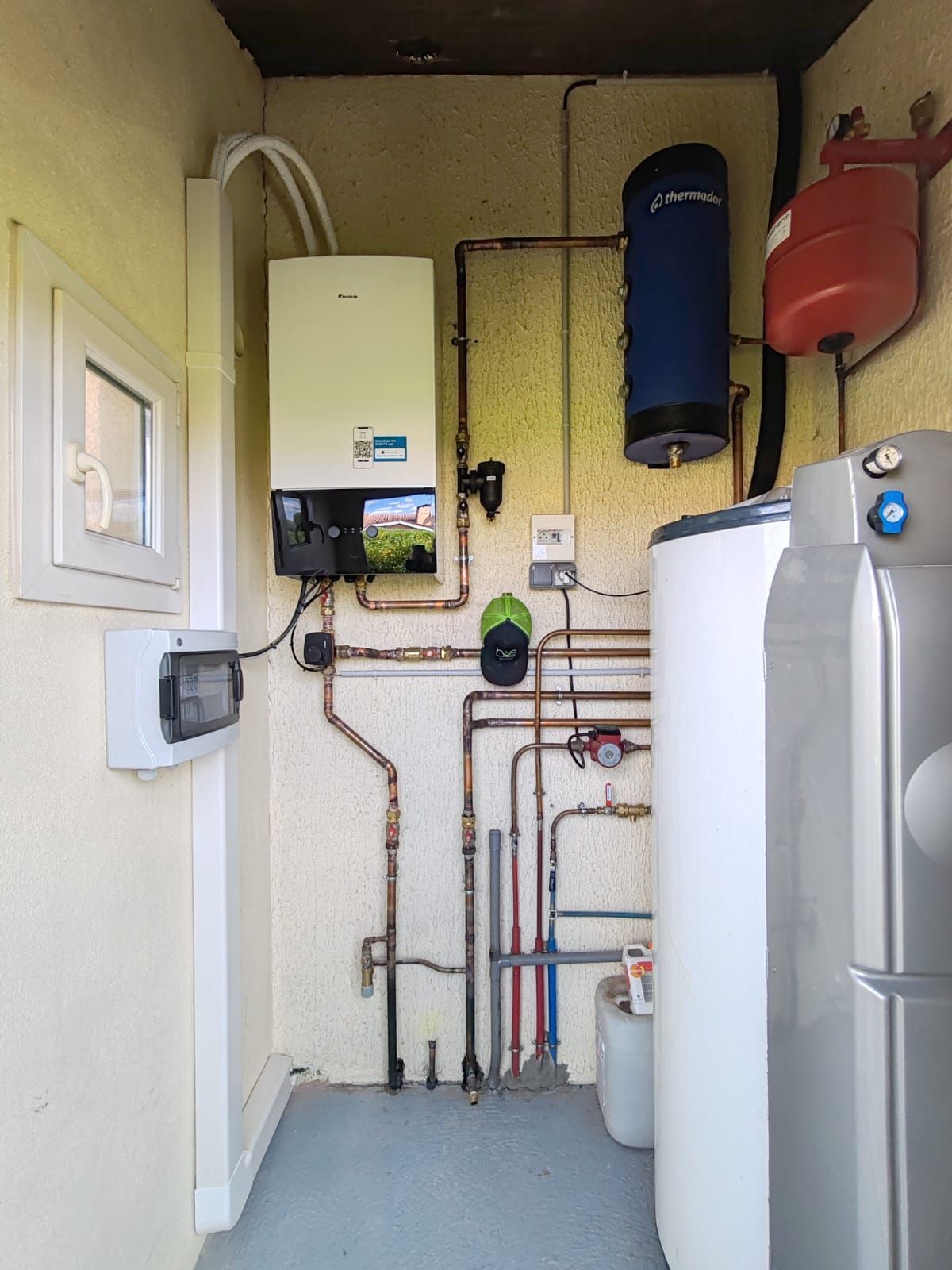 Buanderie avec système de chauffage, tuyaux en cuivre, réservoirs blancs et bleus, petite fenêtre.