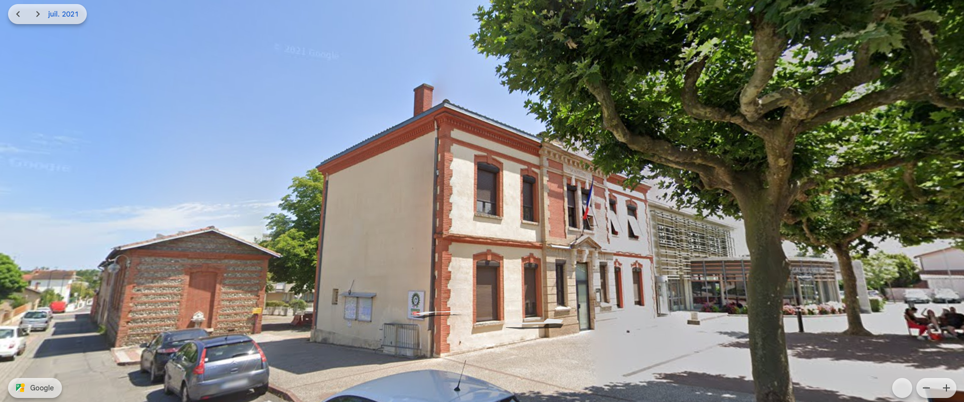Un immeuble de deux étages au toit rouge, entouré d'arbres et de voitures garées. Un ciel bleu s'étend au-dessus.
