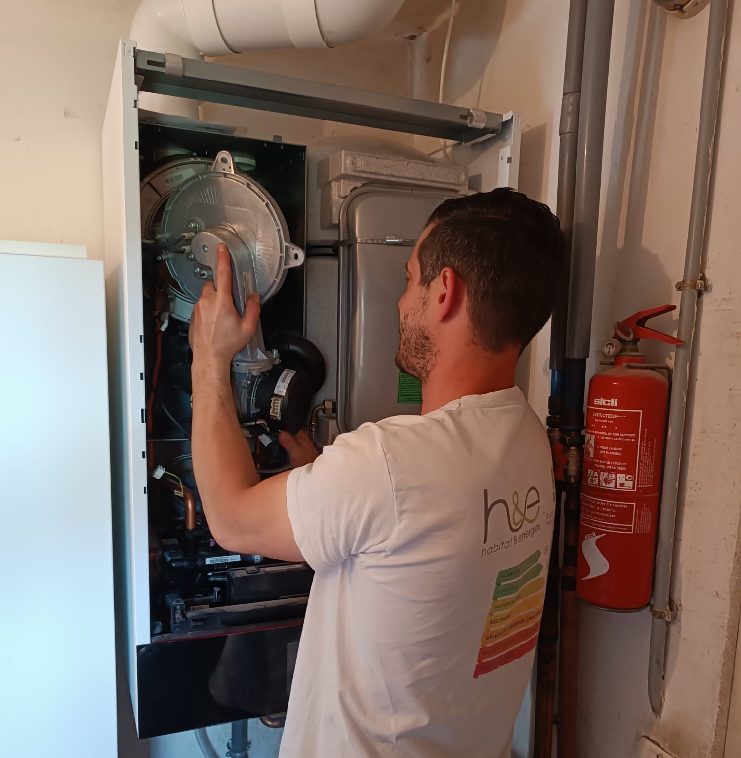 Un homme en chemise blanche répare une chaudière dans une buanderie, à côté d'un extincteur.