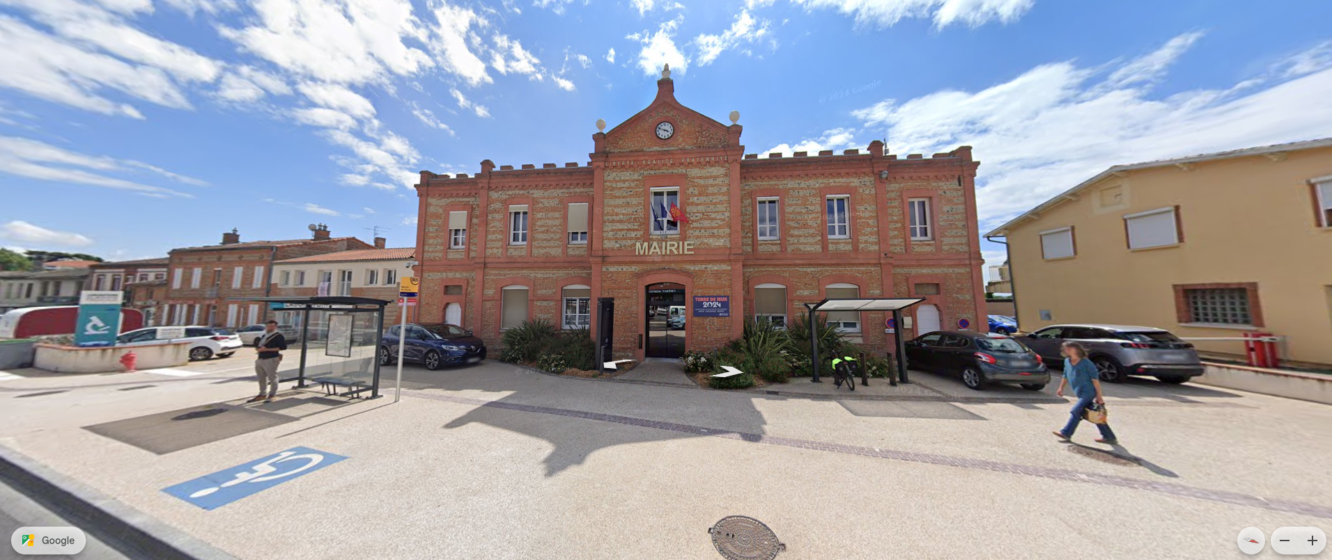 Vue extérieure d'un bâtiment en briques avec un drapeau, par une journée ensoleillée, avec des gens marchant et des voitures garées dans la rue.
