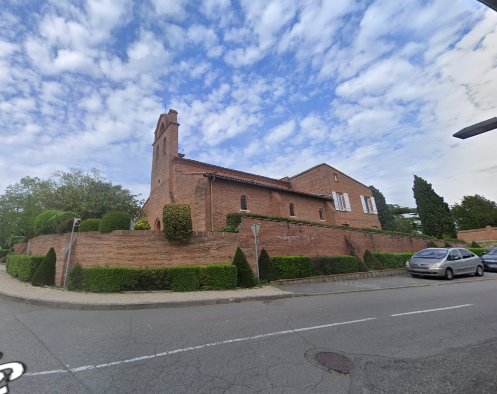 Église en briques avec toit de tuiles rouges et petite tour, à côté d'une route avec des voitures, sous un ciel partiellement nuageux.