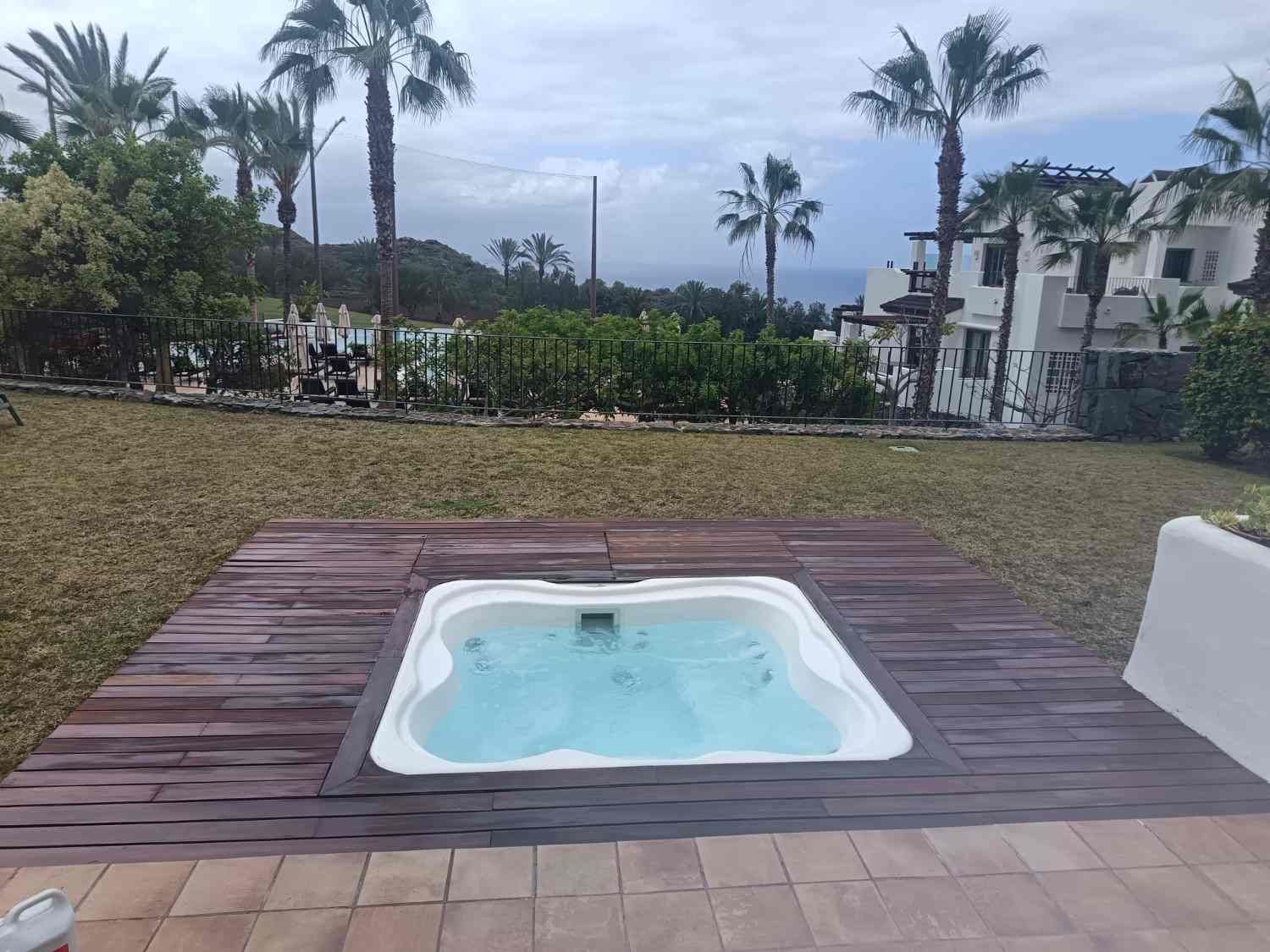 Pequeña bañera de hidromasaje cuadrada sobre una terraza de madera con palmeras y edificios al fondo.