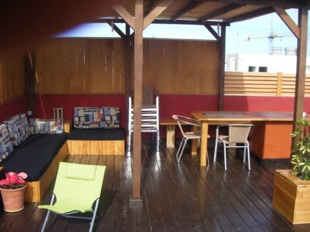 Terraza exterior de madera con bancos, mesa y sillas bajo un techo cubierto, con barandillas rojas.