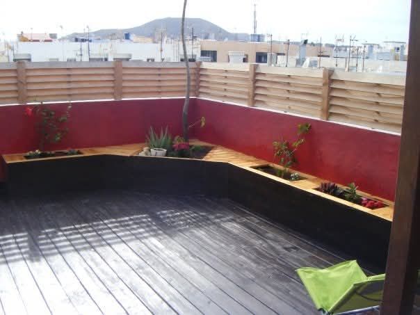 Terraza vacía en la azotea con paredes rojas, suelo de madera oscura y valla de madera que proporciona privacidad, con vistas a colinas lejanas.