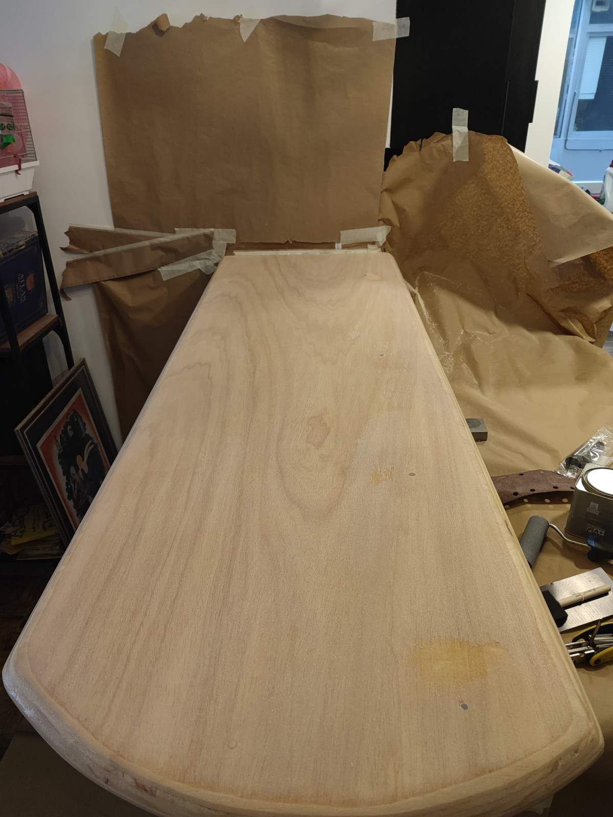 Tabla de madera sin acabado sobre un banco de trabajo, con cartón y herramientas en un taller.