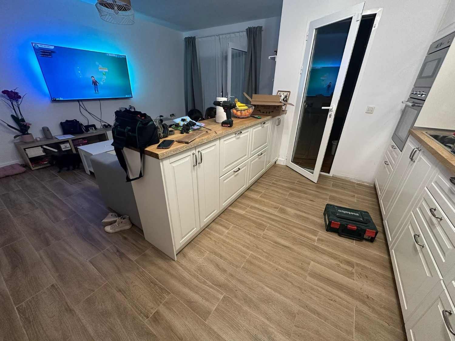 Cocina moderna con armarios blancos, suelo de madera, cuadros con iluminación azul en la pared y una puerta que da a un balcón oscuro.